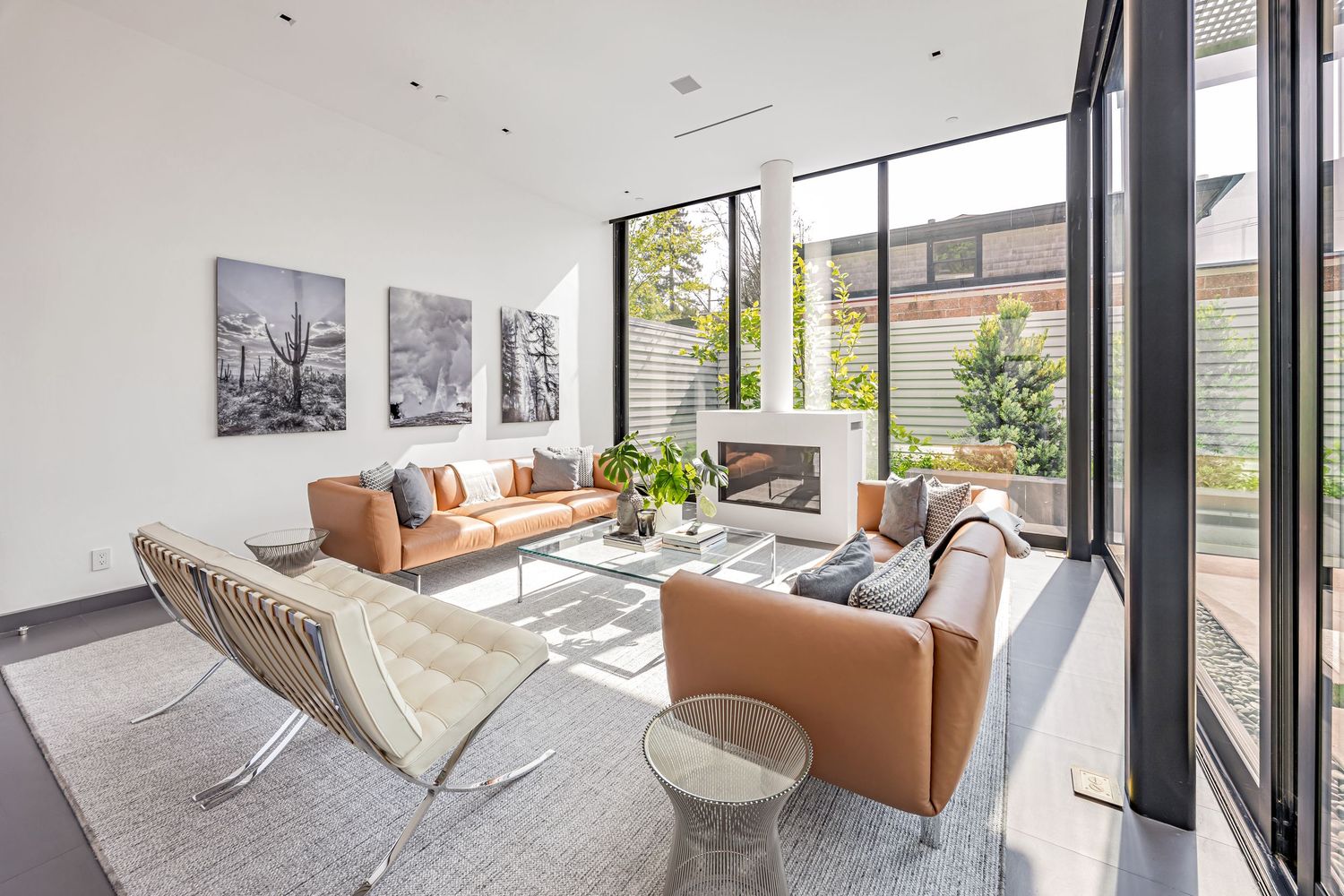 Sunlit living room with tan leather furniture, geometric rug, and floor-to-ceiling windows overlooking garden.
