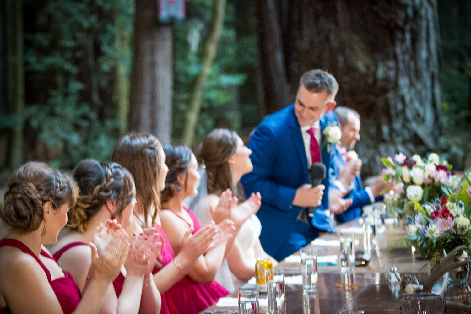 Wedding party in pink dresses applauding during outdoor forest reception.
