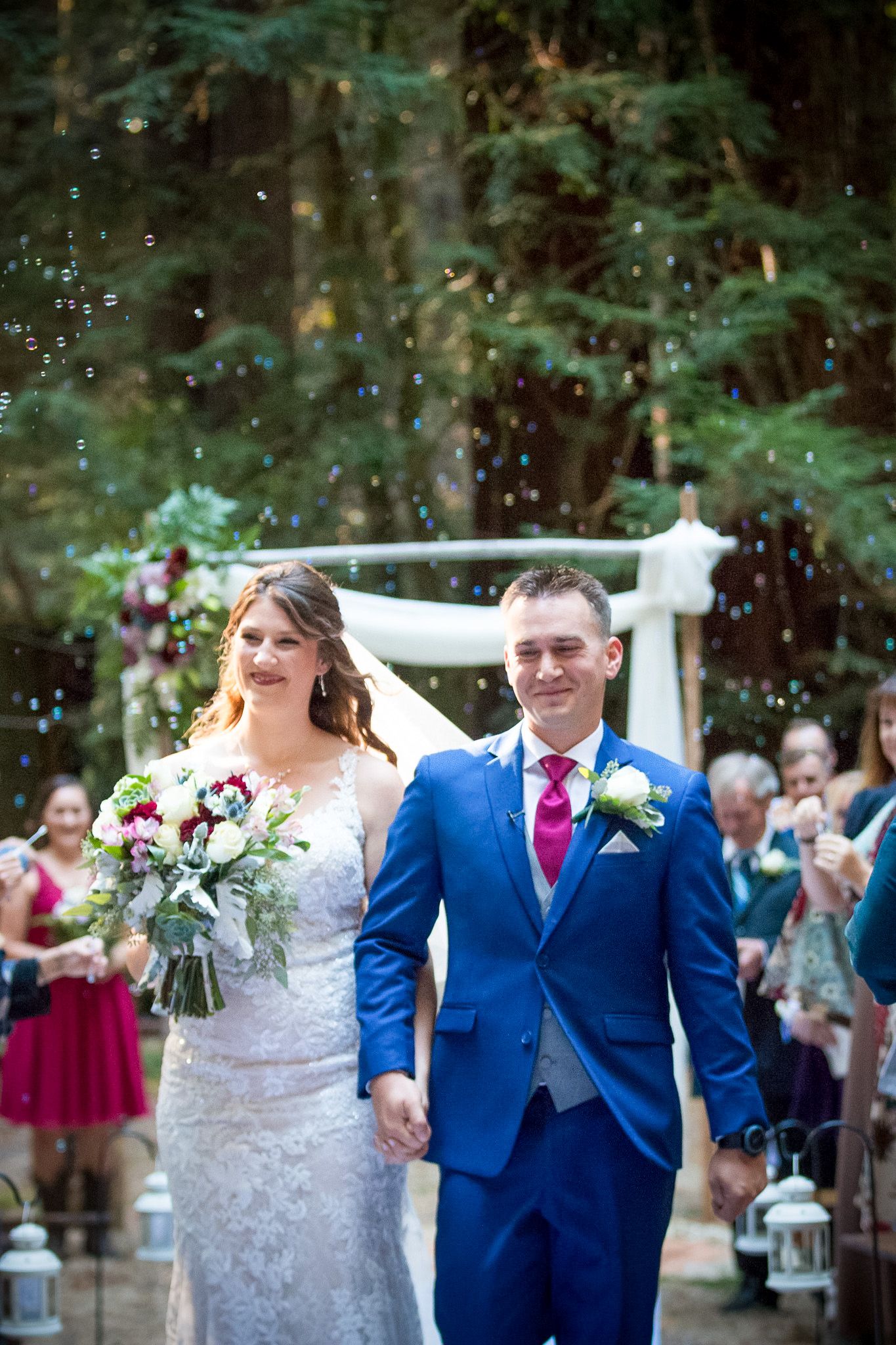 Joyful couple exits their forest wedding ceremony as guests shower them with bubbles.
