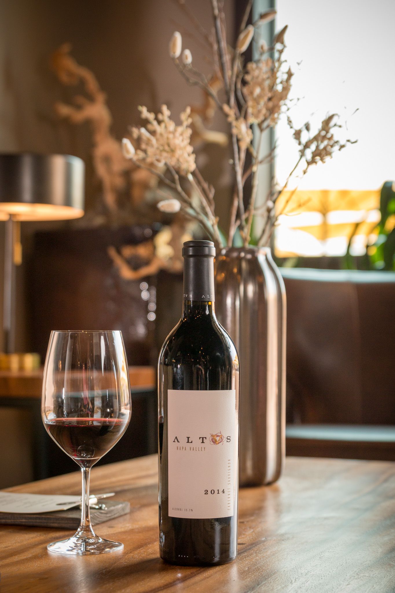 Alter wine bottle and filled glass on wooden table with dried floral arrangement in background.