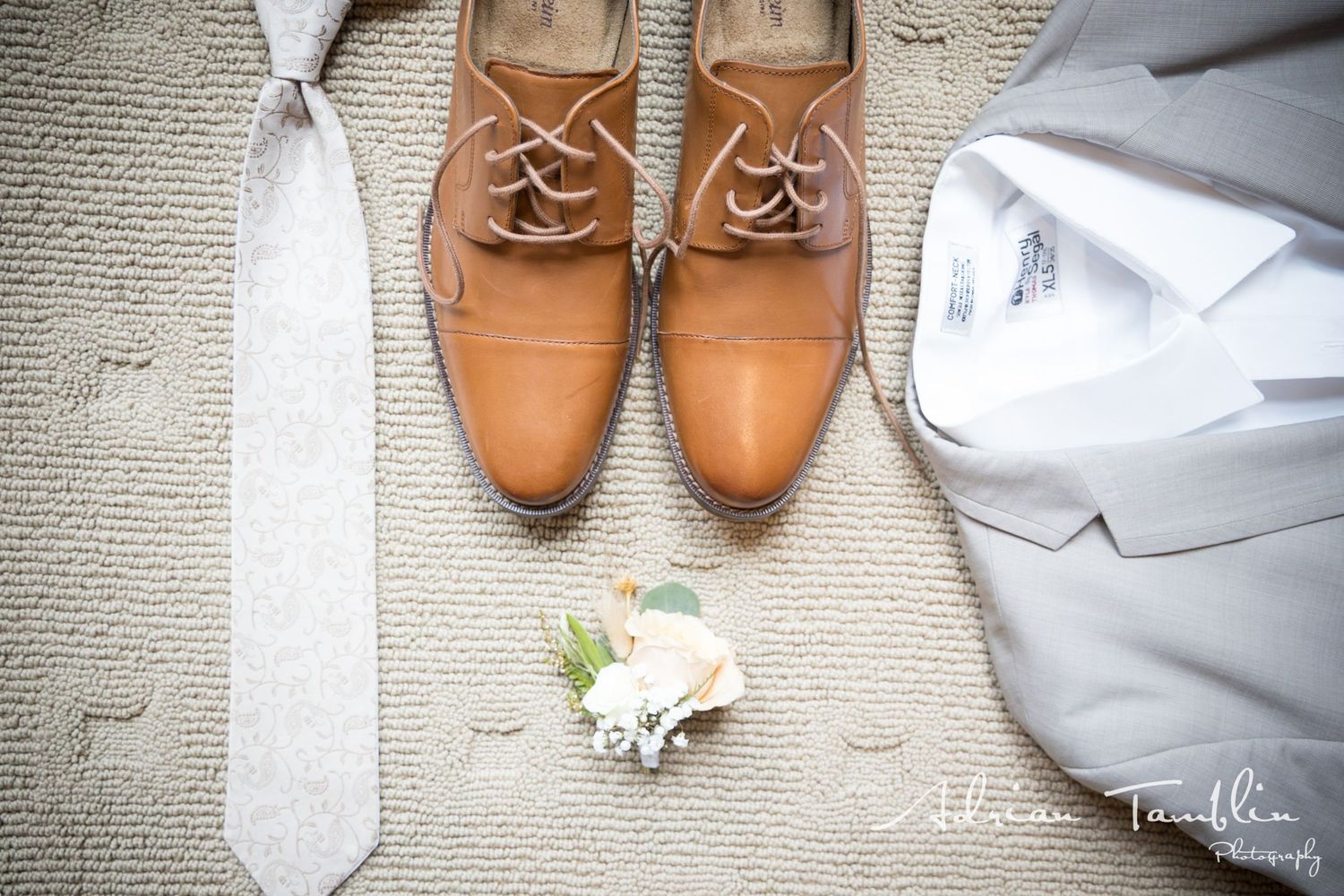 Brown leather dress shoes with tie and boutonniere arranged on beige carpet.