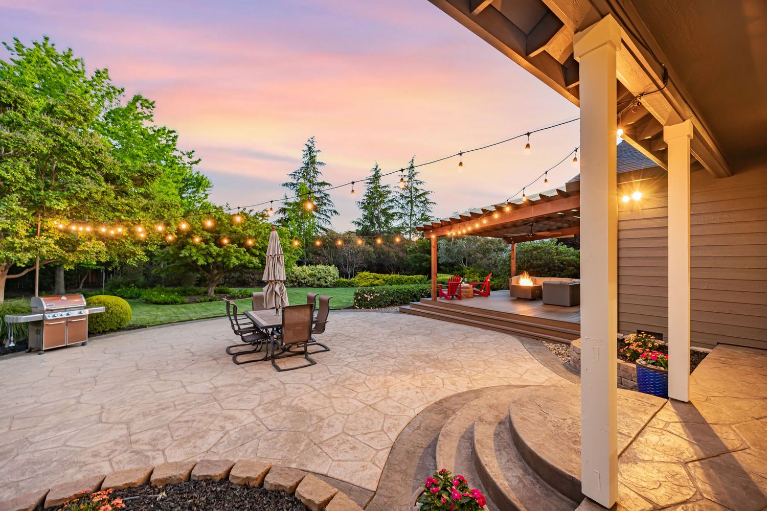 Elegant curved patio with string lights and fire pit area against a colorful sunset backdrop.