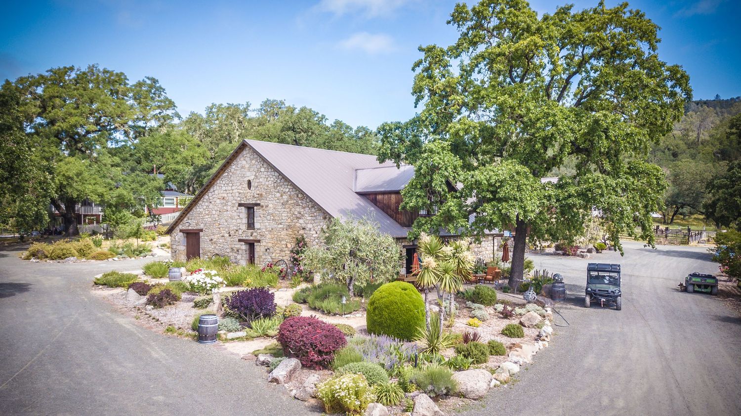 Rustic stone winery building with landscaped gardens and a circular driveway nestled among lush green trees.