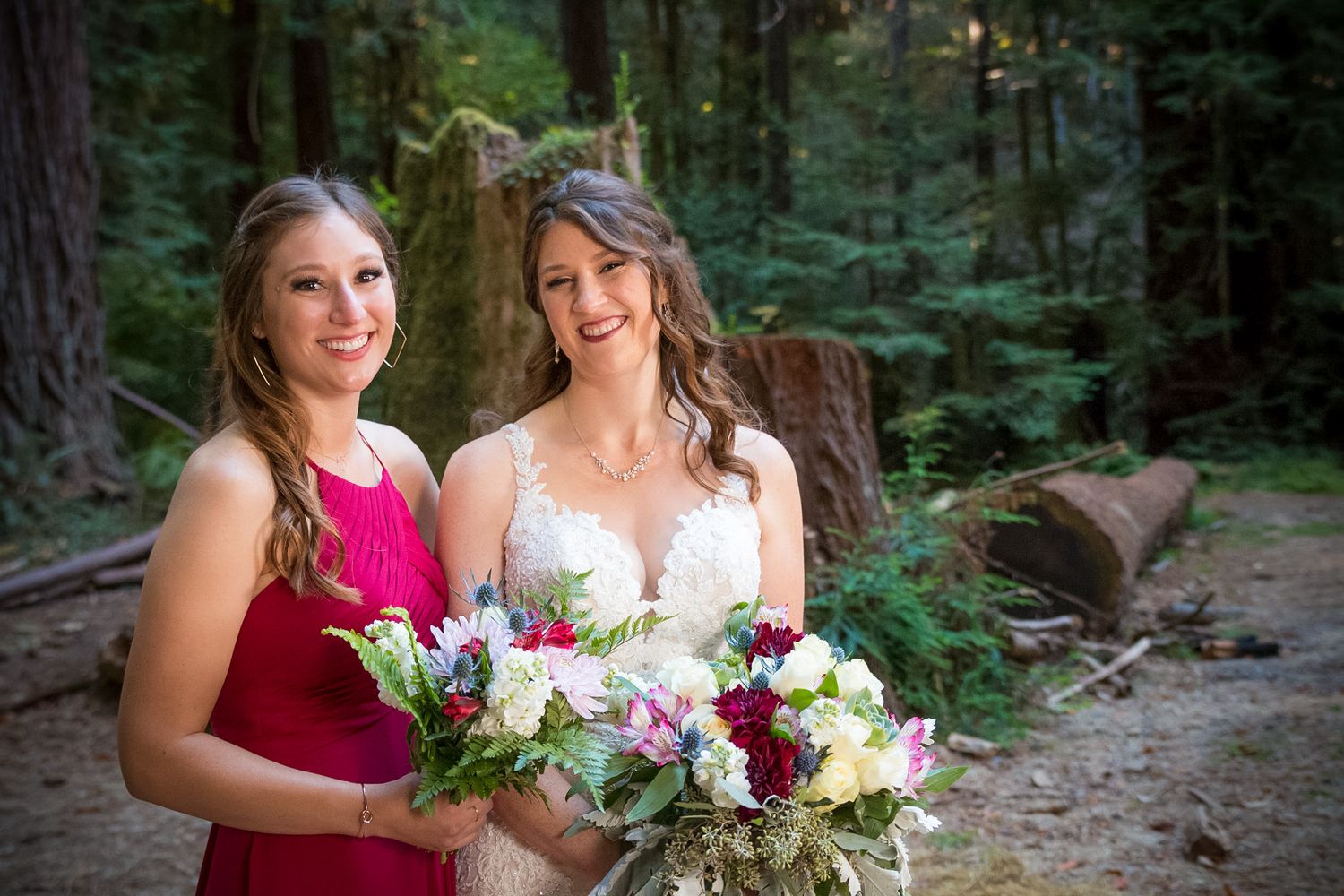 Bride and bridesmaid share joyful moment while holding floral arrangements in wooded setting.