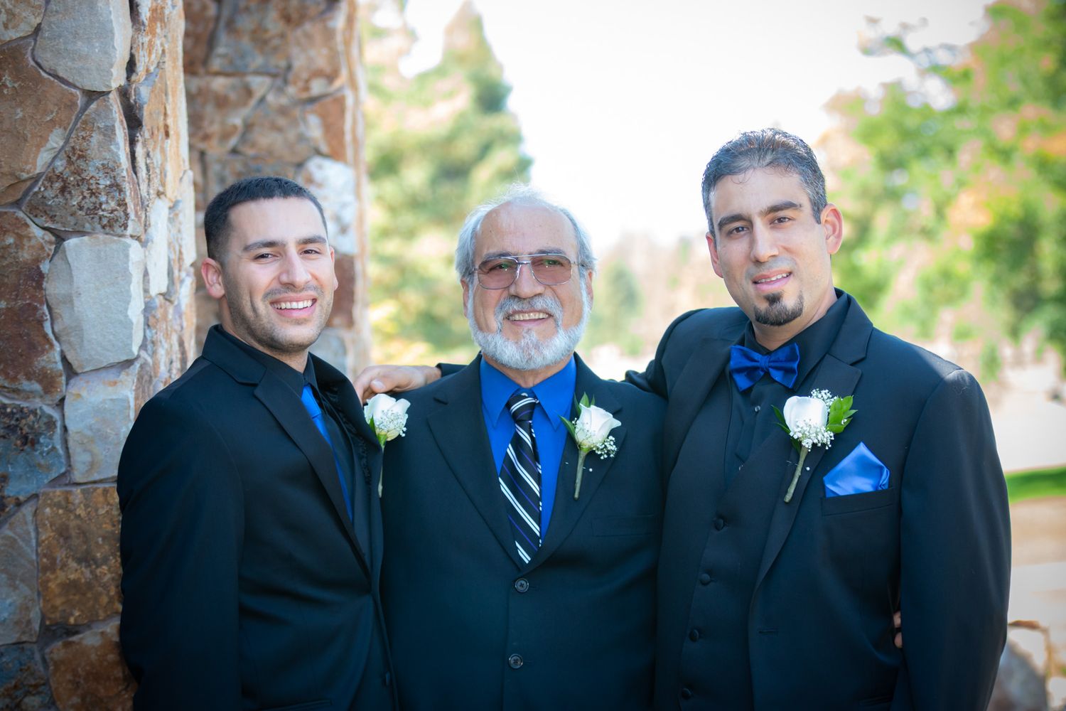 Three men in formal suits with boutonnieres pose together at a wedding.
