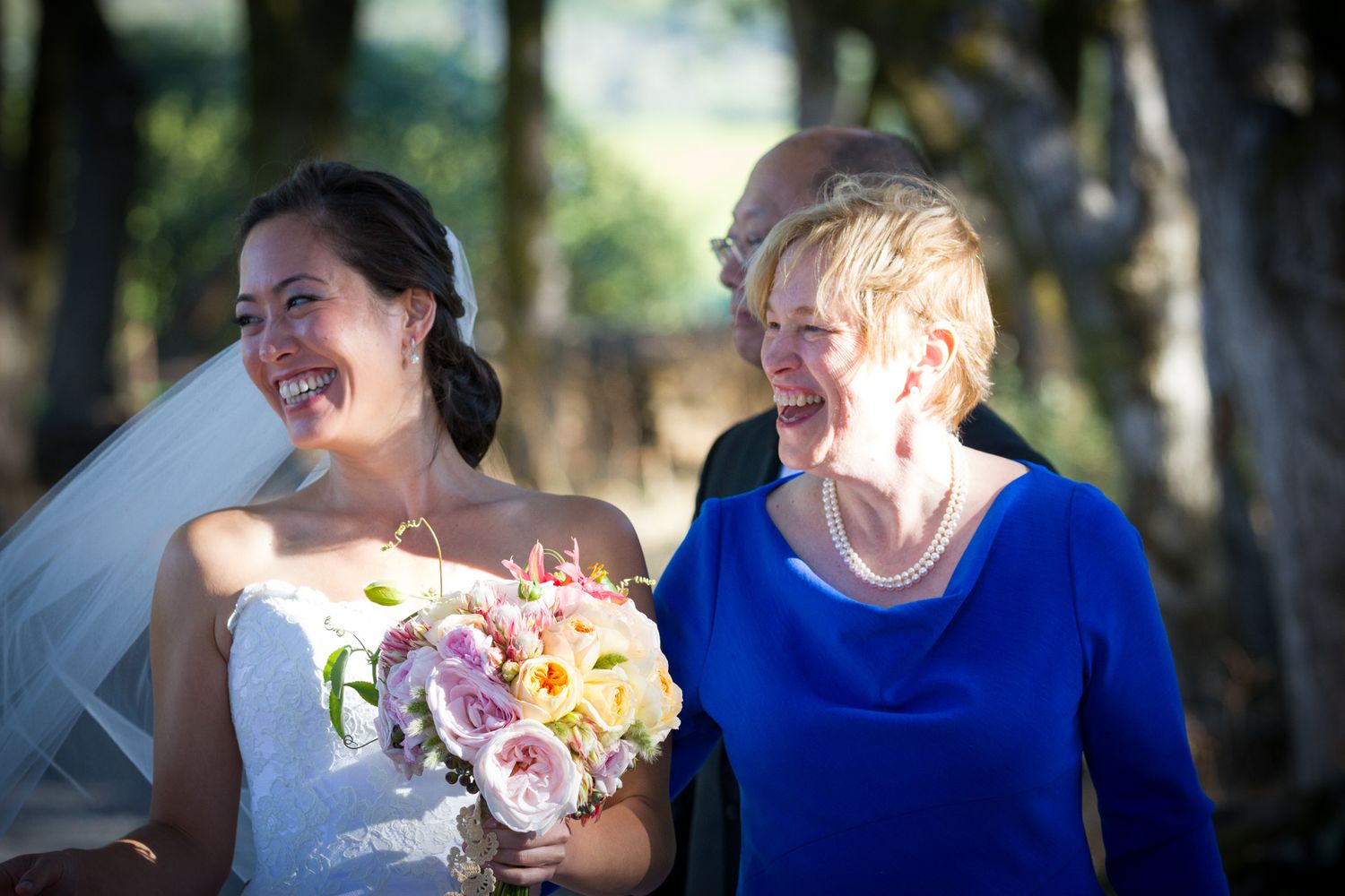 Two wedding attendants sharing joyful laughter while holding bridal bouquet outdoors.