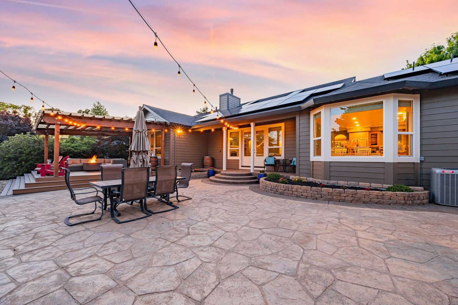 Contemporary backyard features stone patio with string lights and comfortable seating under pink sunset sky.