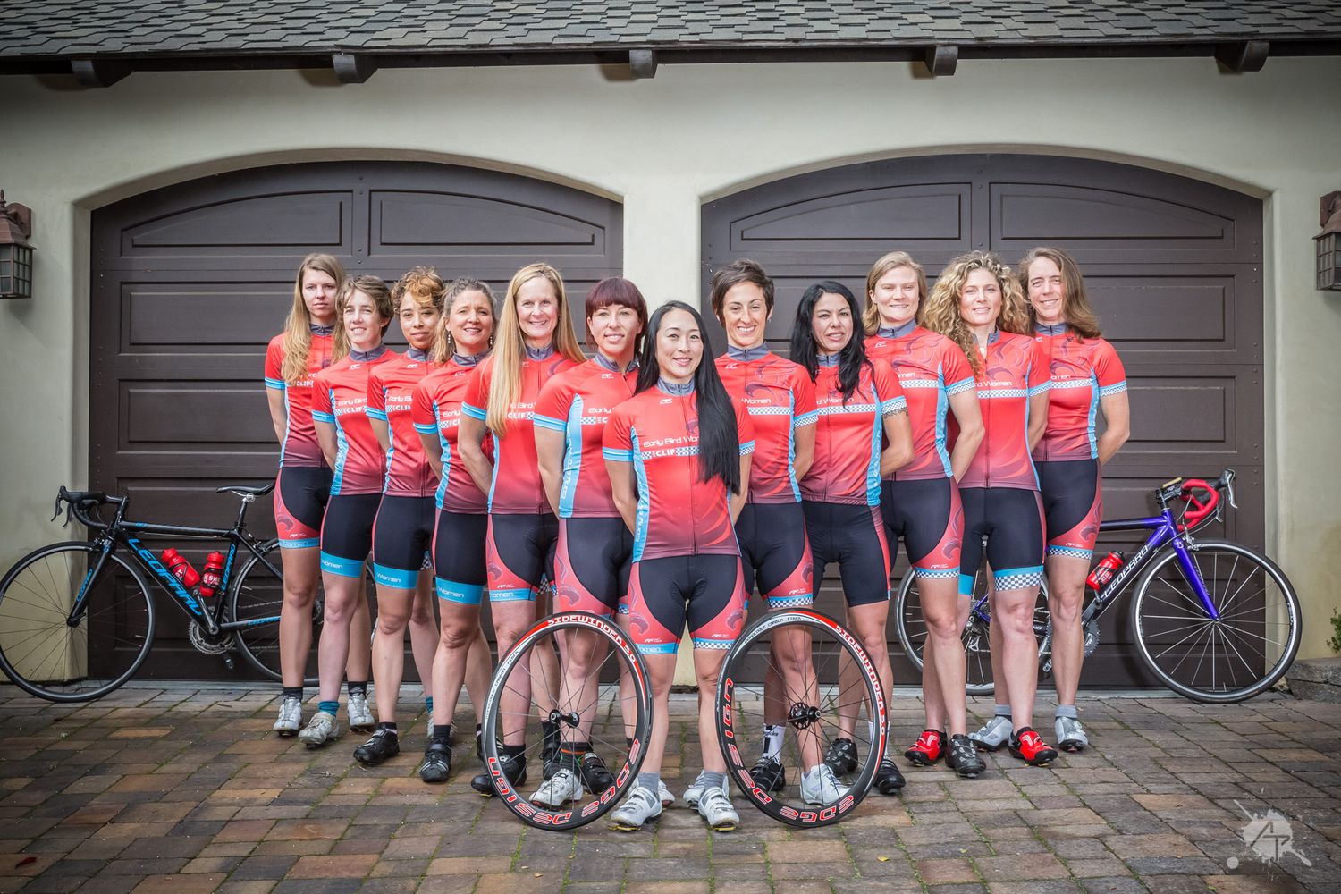 Group of cyclists in matching red jerseys and grey shorts pose with their bikes in front of garage doors.