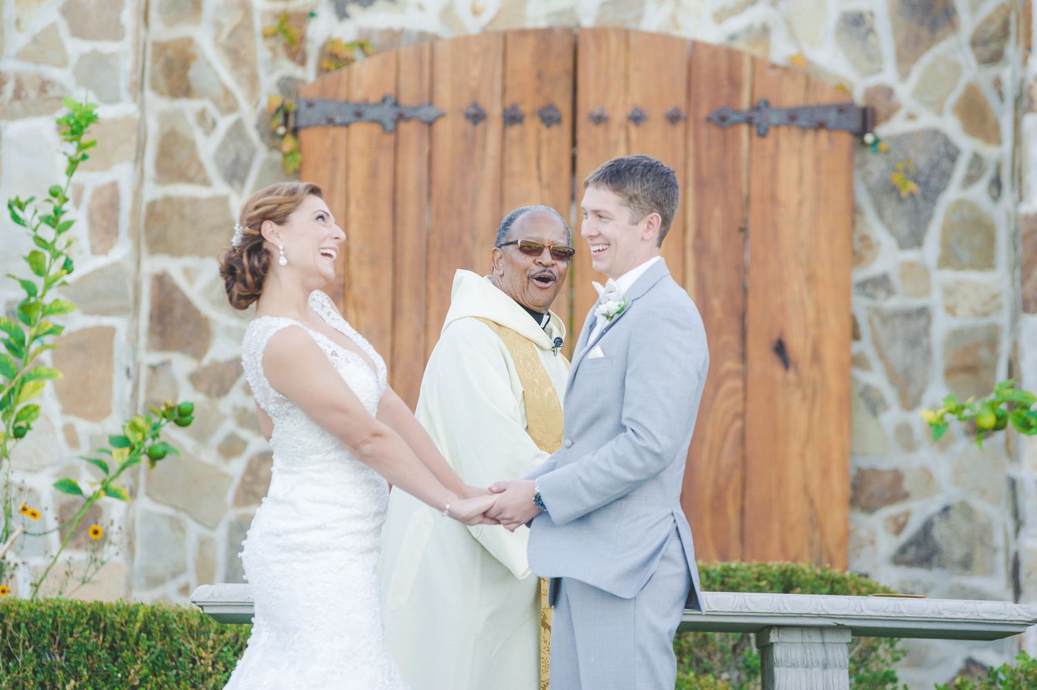 A couple holds hands during an outdoor wedding ceremony in front of a rustic wooden door and stone wall.