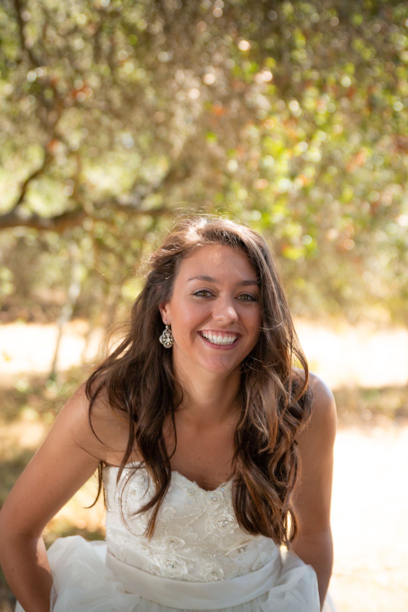 A bride in a white lace dress smiles brightly against a soft, sun-dappled outdoor background.