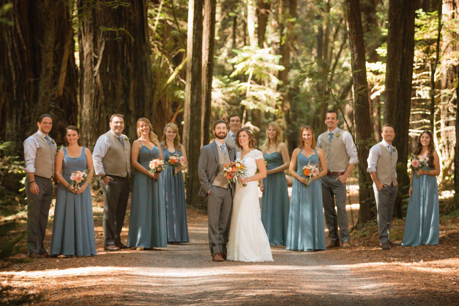 Wedding party gathered on forest path with bridesmaids in blue dresses and groomsmen in gray.
