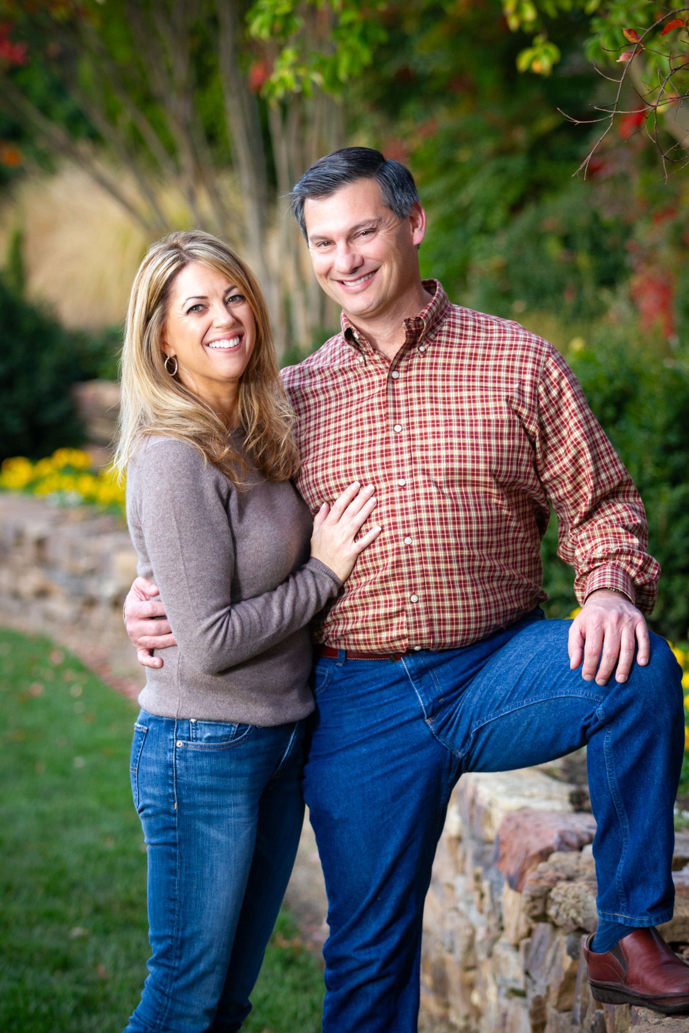 Casual outdoor portrait of couple in coordinating outfits against stone wall and fall foliage.