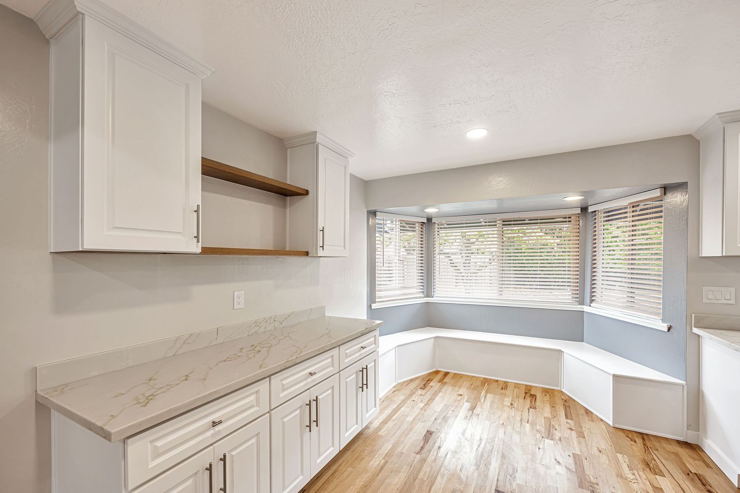 Bright kitchen interior with white cabinets, curved window seat, and wood flooring.