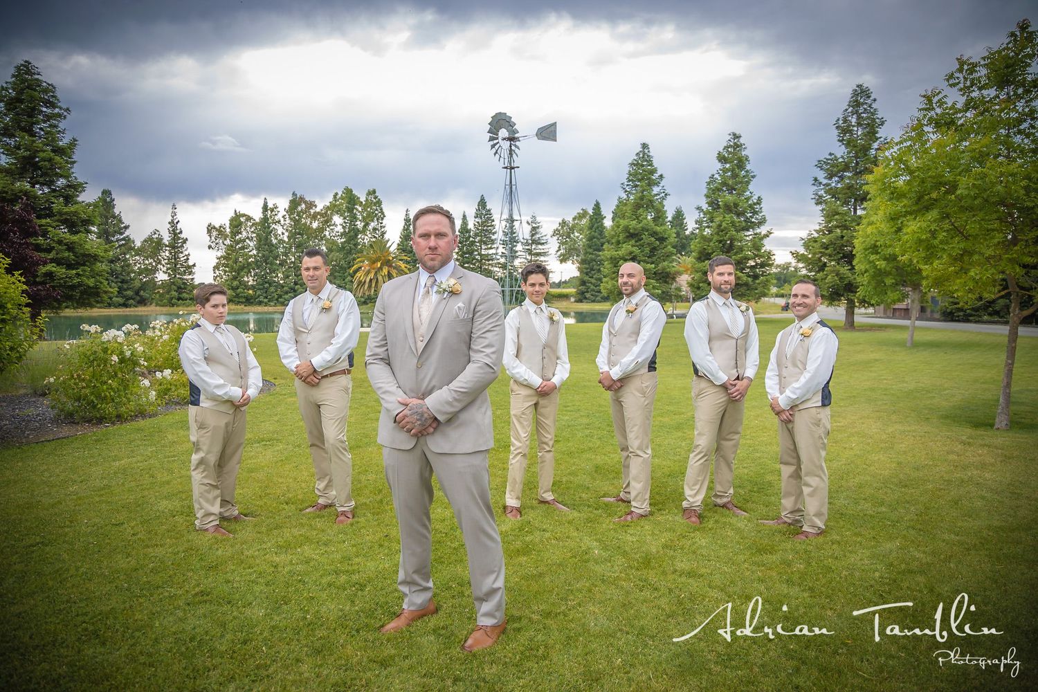A wedding party in light gray suits poses outdoors under dramatic storm clouds near a windmill.