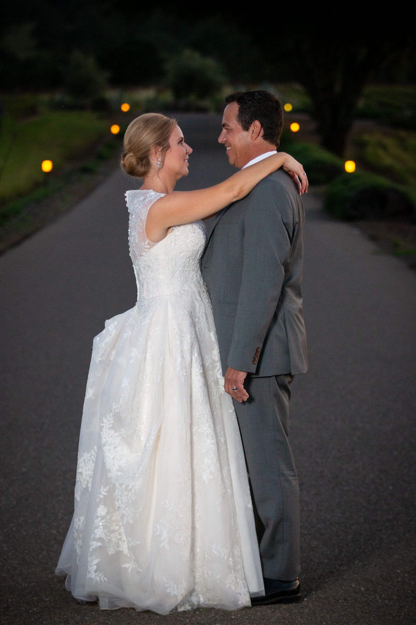 Romantic evening portrait of newlyweds embracing on path with glowing lights in background.