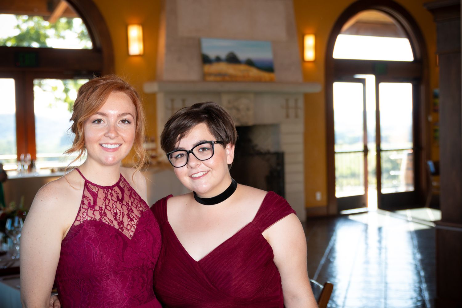 Two people in burgundy dresses smile together in elegant venue with arched windows.