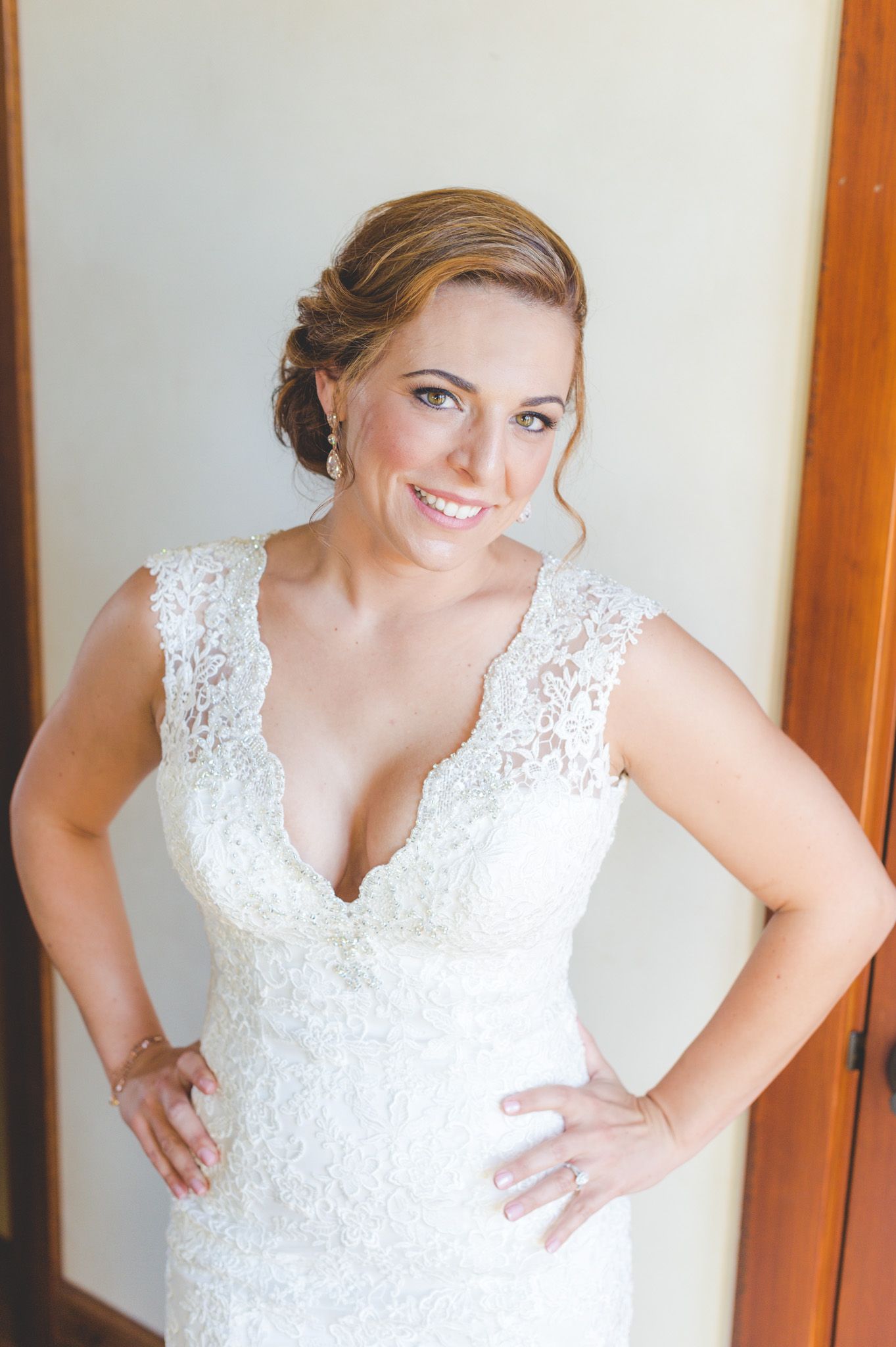 A bride poses in a white lace wedding dress with v-neckline and delicate straps against a light background.