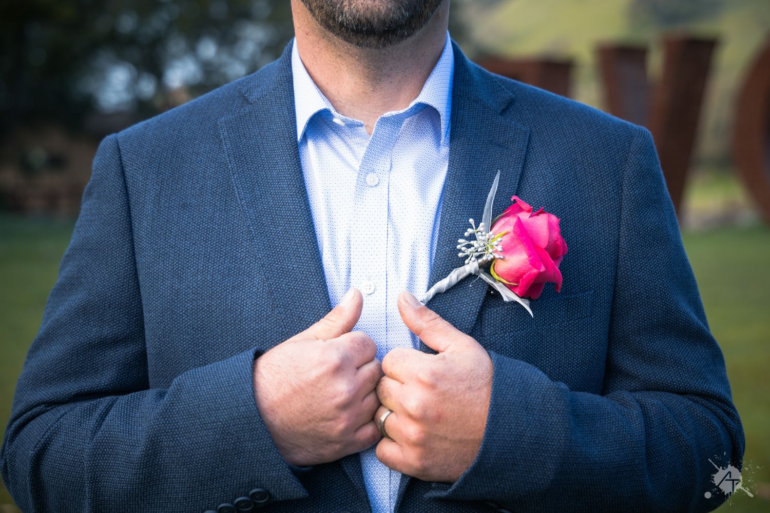 A groom in a dark blue suit adjusts his lapel while wearing a vibrant pink rose boutonniere.