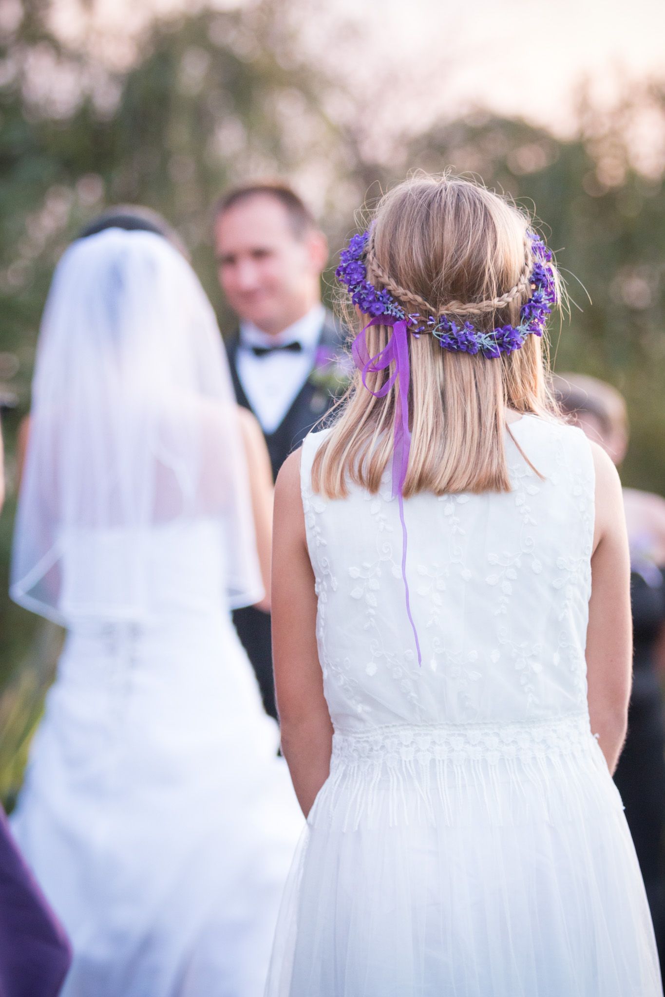 Row of bridesmaids in white dresses with purple floral crowns shown from behind at outdoor wedding ceremony.