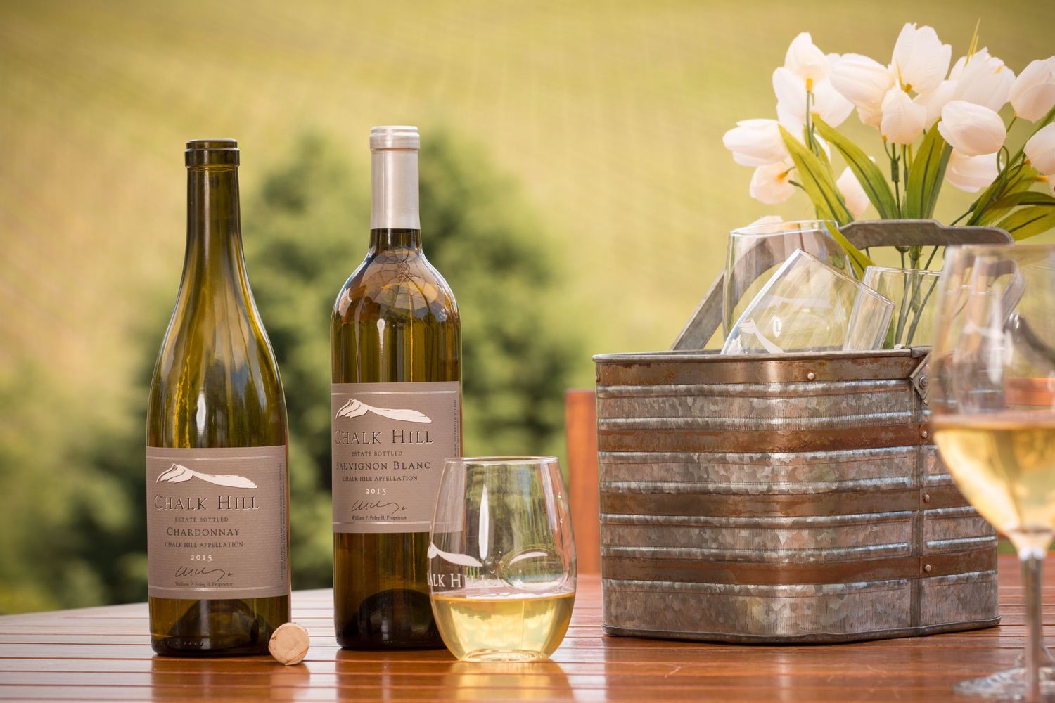 Two wine bottles and glass displayed with white tulips and stacked wooden boxes.
