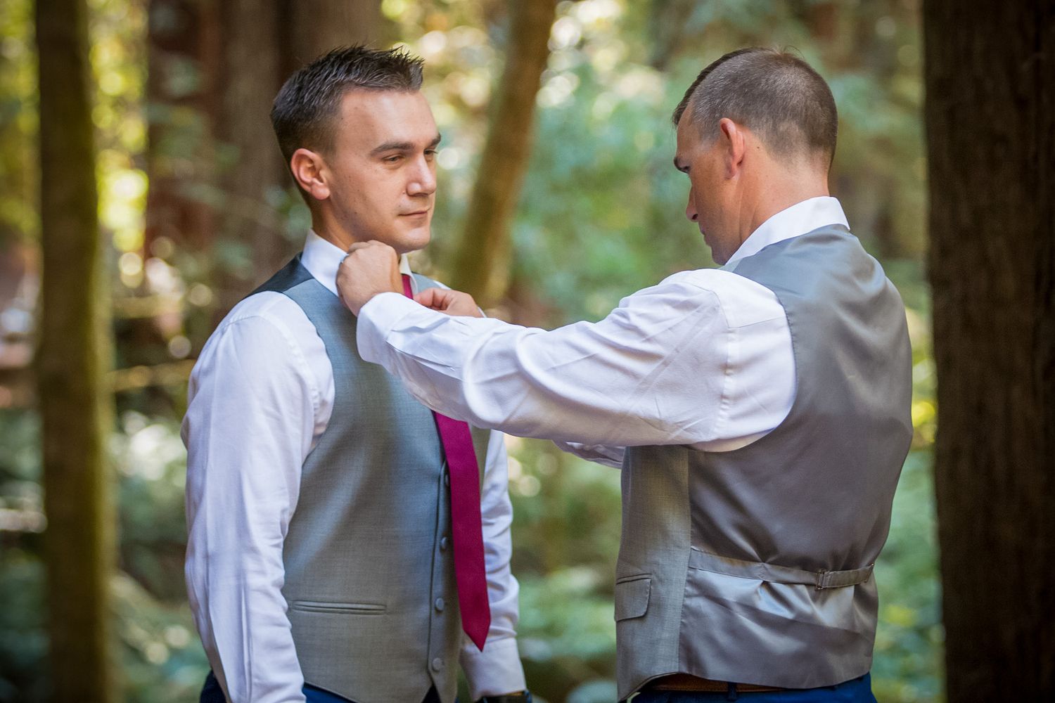 Groomsman adjusts another's burgundy tie before outdoor forest wedding ceremony.