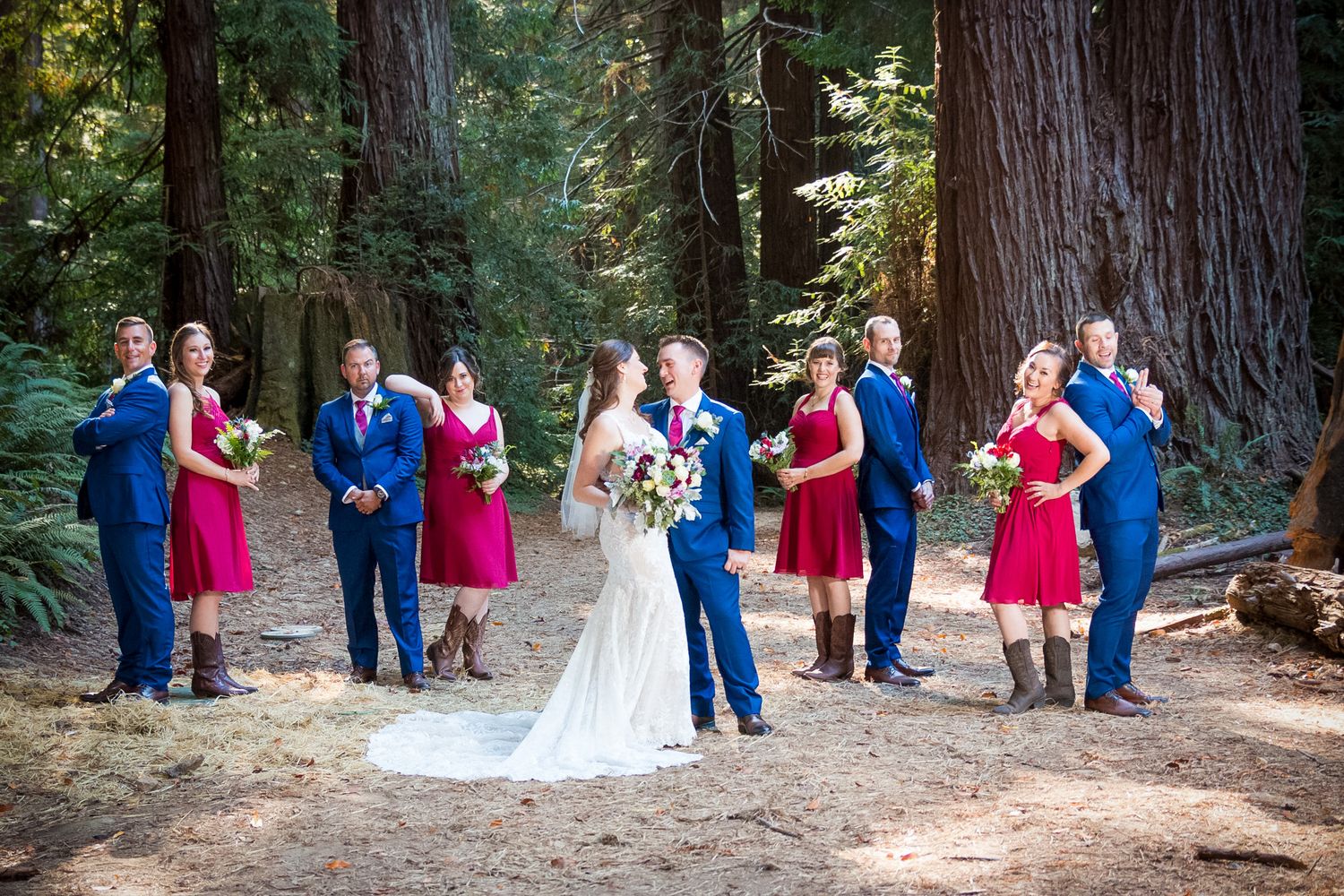 Wedding party in coordinated red and blue attire poses for formal photos in a redwood forest.