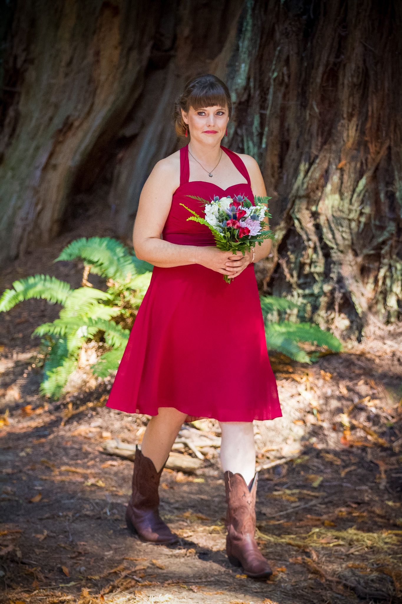 Bridesmaid in red dress and cowboy boots holding colorful bouquet against redwood tree.