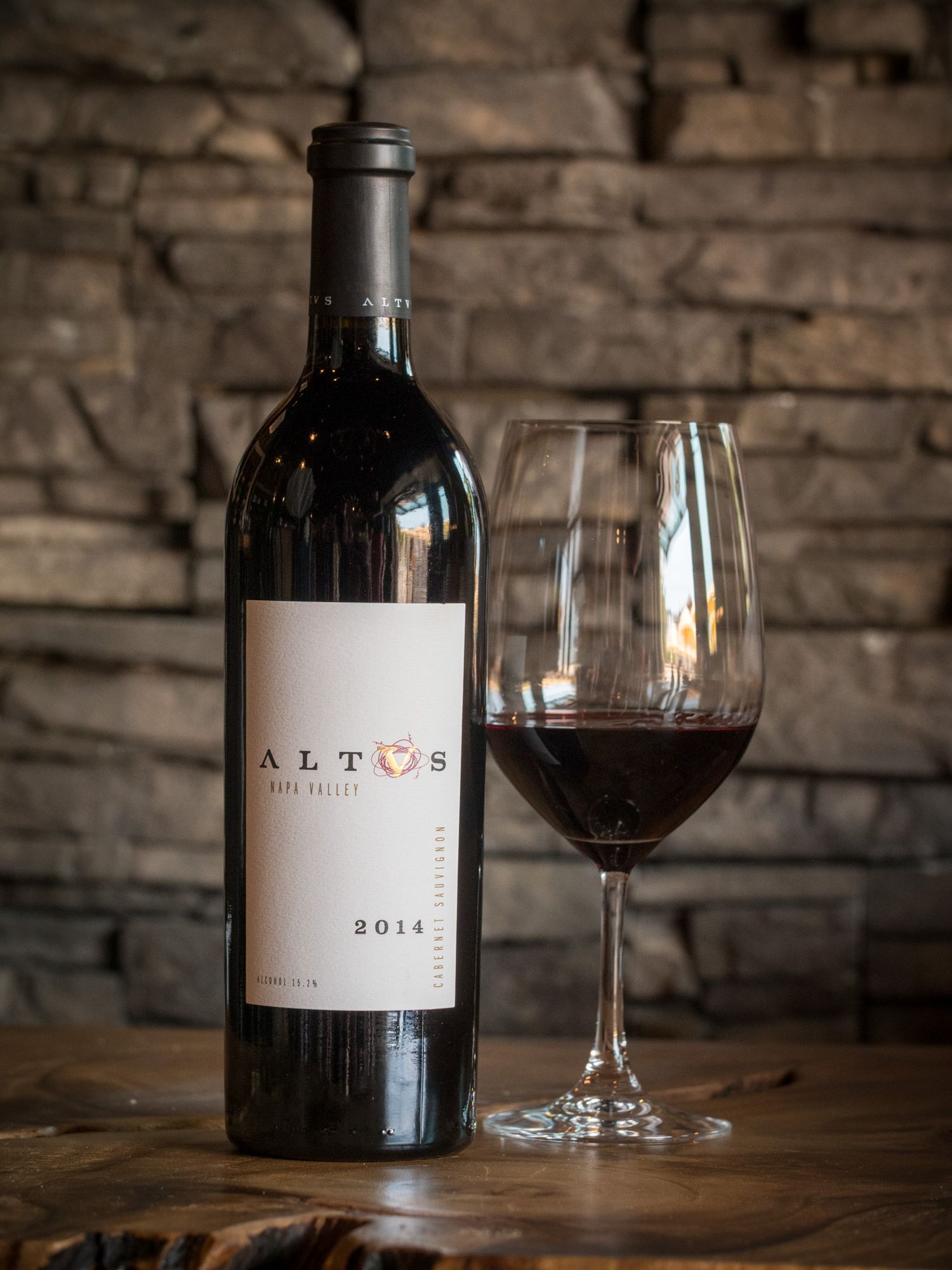 Altus wine bottle and glass of red wine against rustic stone wall background.