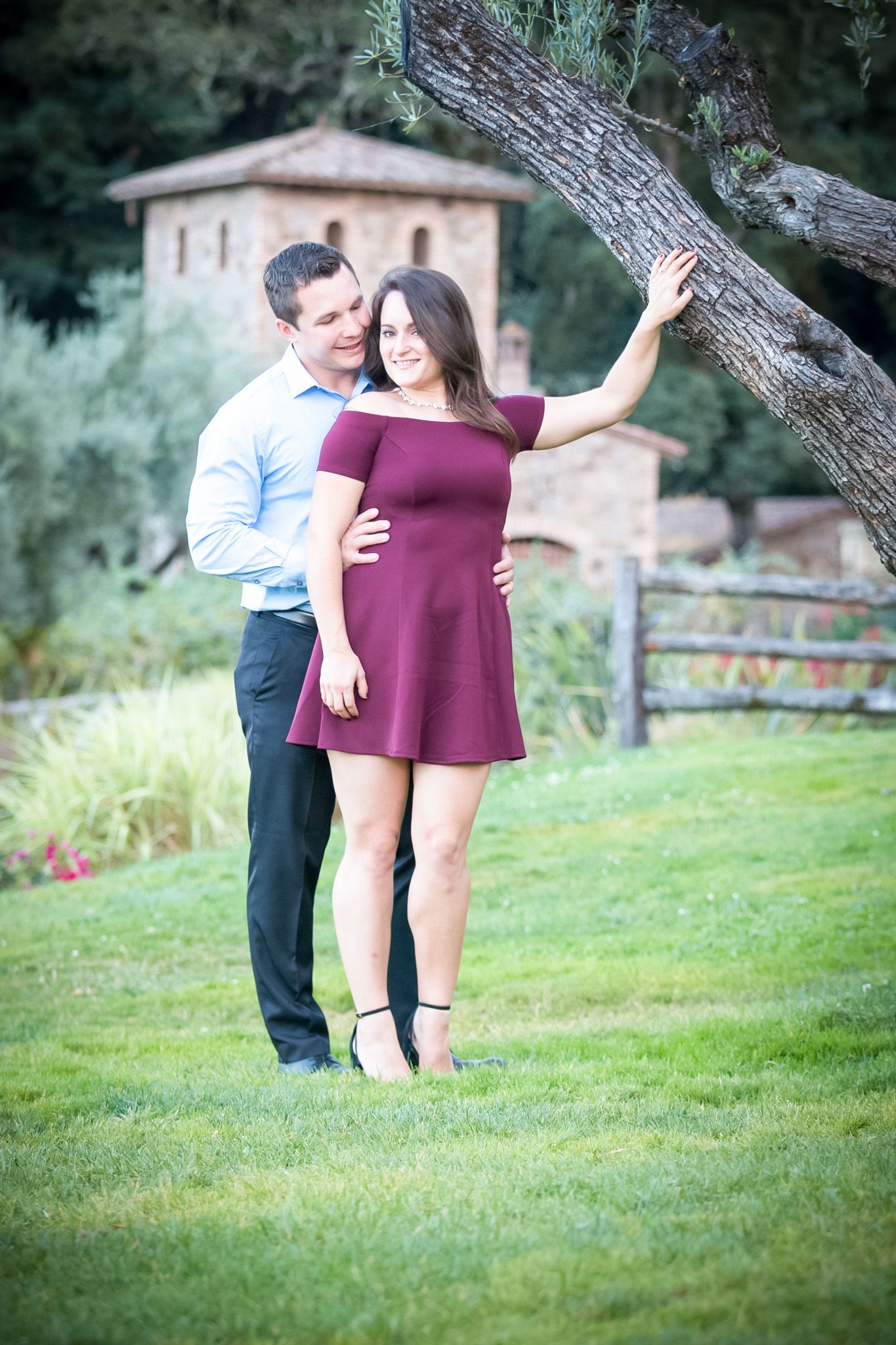 A couple poses together outdoors near a stone building, with the woman in a burgundy dress leaning against a tree.
