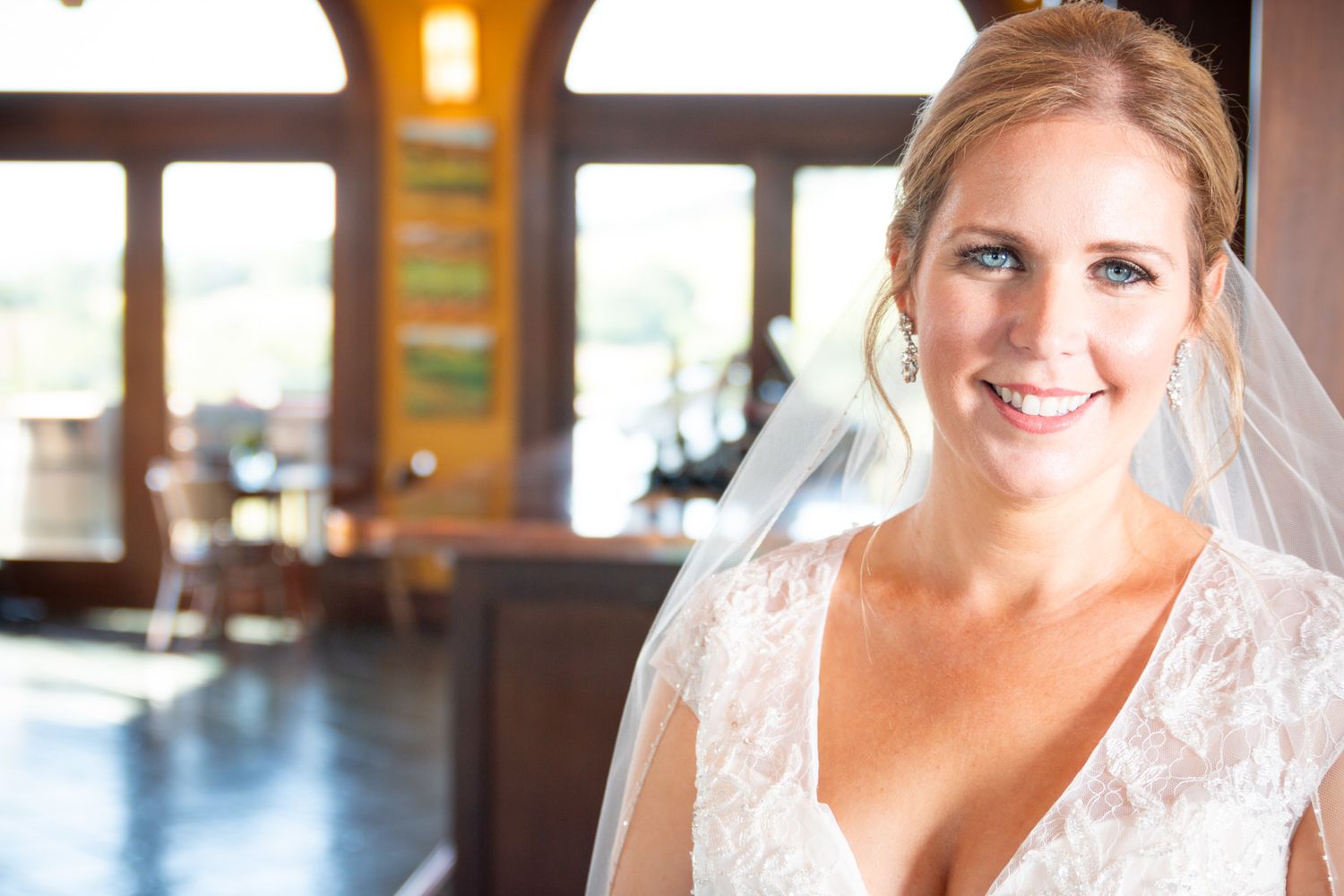 A bride in a white lace gown poses inside a venue with warm lighting and wooden details.