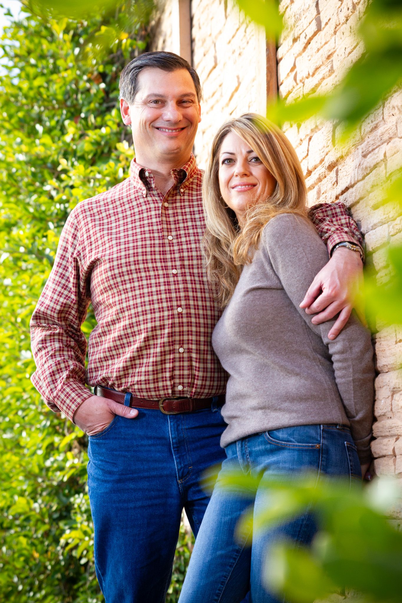 Happy couple embracing against stone wall with sunlit greenery in background.