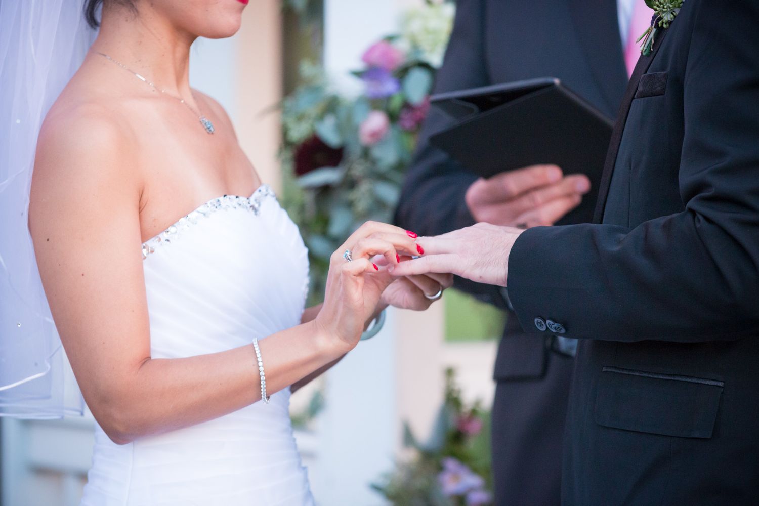 Exchange of wedding rings during ceremony with purple floral arrangements in background.
