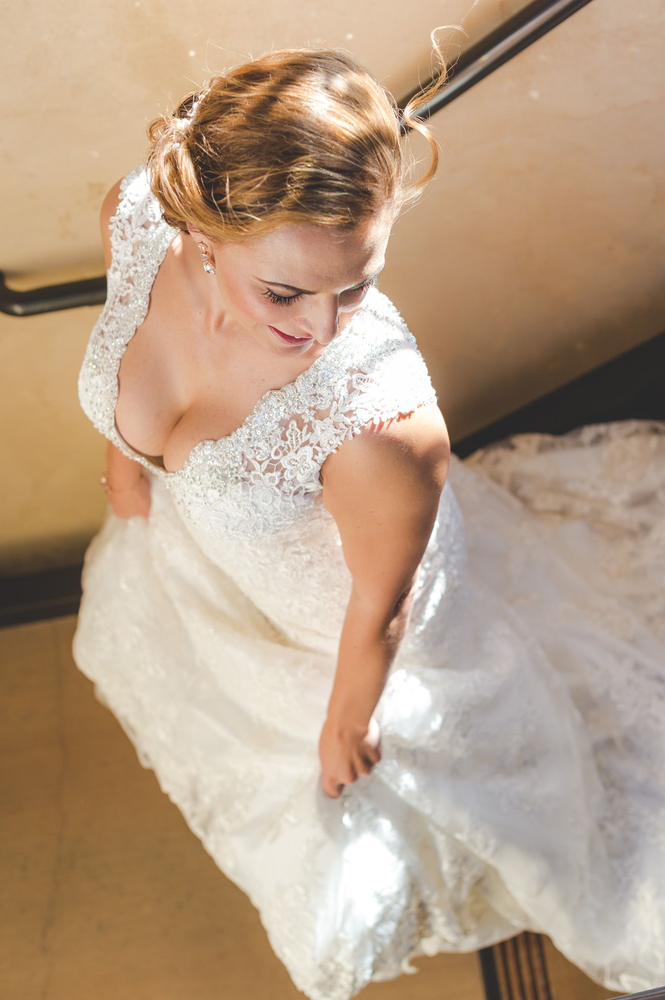 A bride in an elegant lace wedding dress with cap sleeves looks down while standing.