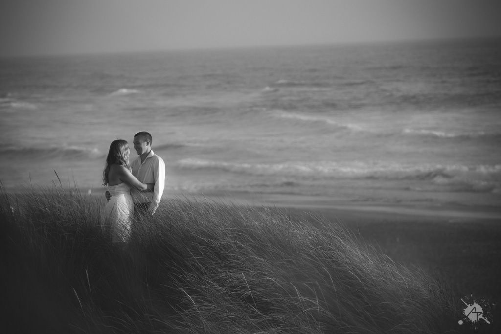 A couple embraces in tall grass on a dramatic coastal overlook with moody ocean waves in black and white.