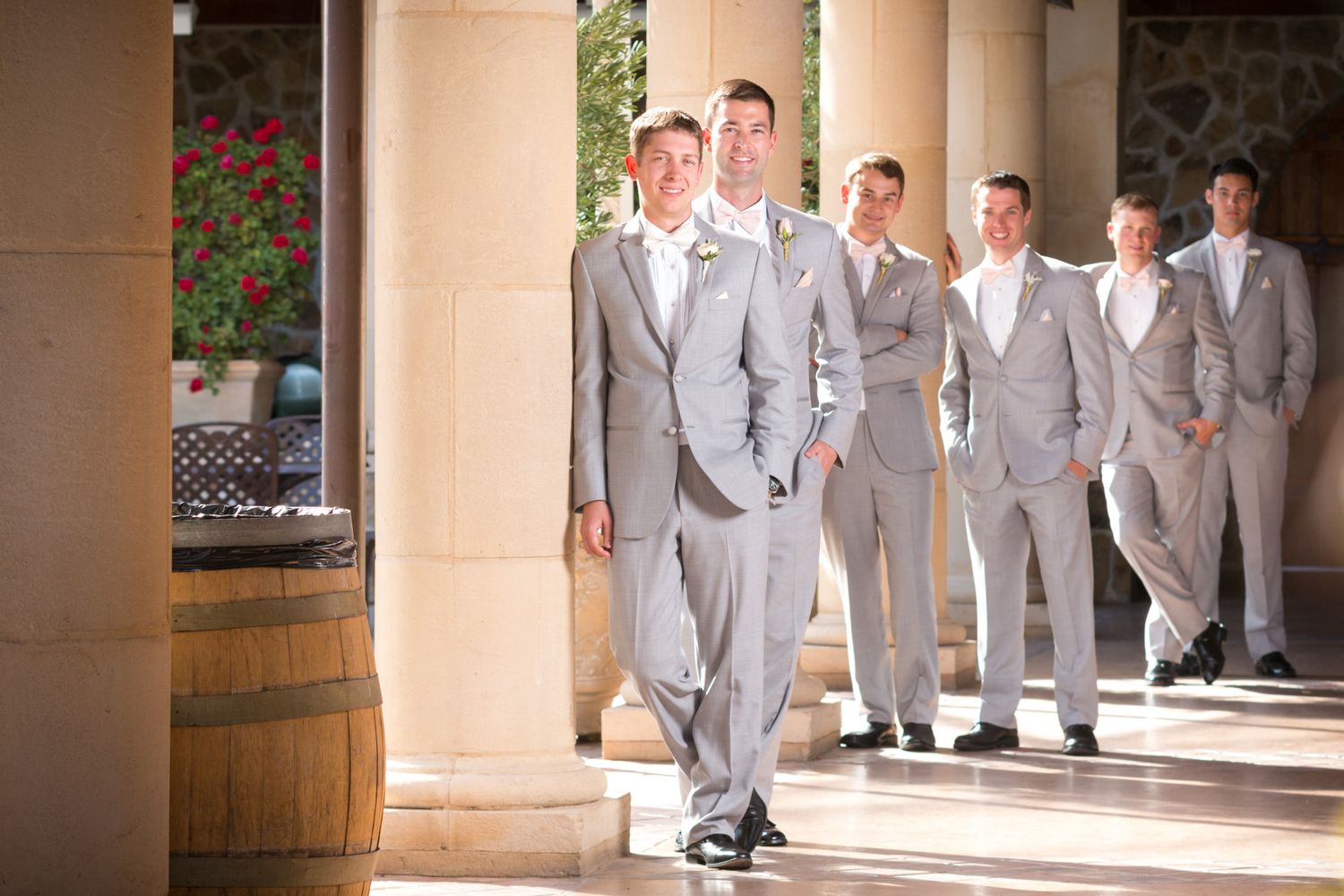 Groomsmen in light gray suits walking through a Mediterranean-style colonnade.