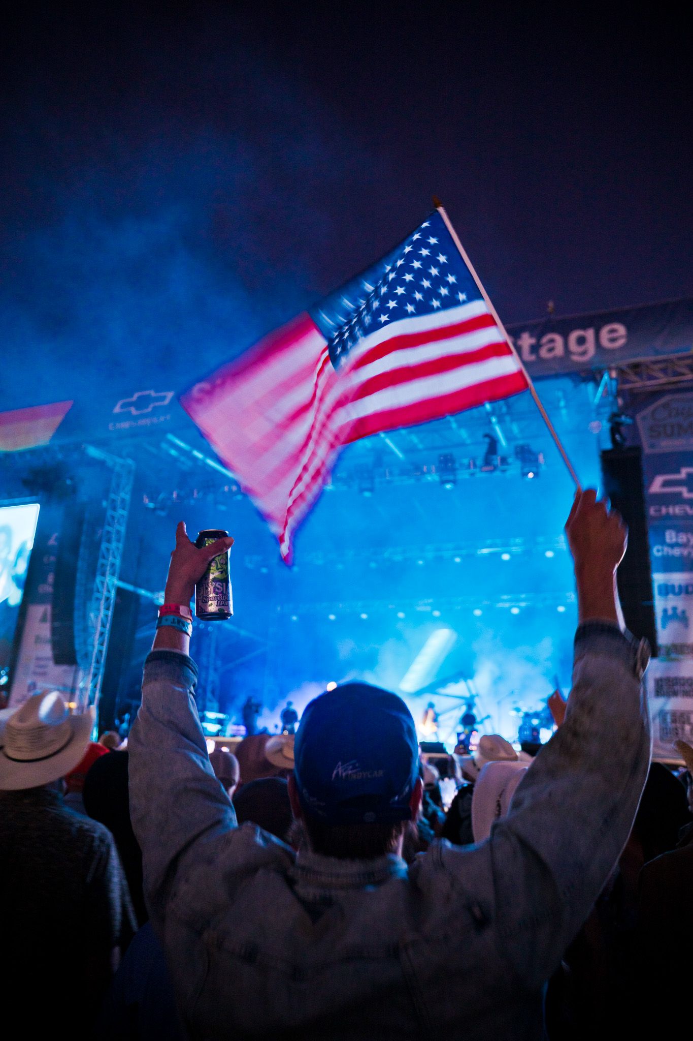 Concert crowd member waves American flag in front of illuminated blue stage at outdoor music festival.