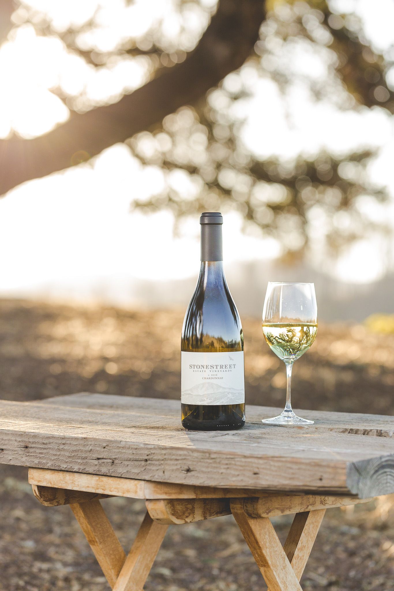 White wine bottle and glass on rustic wooden table with golden sunlit trees in background.