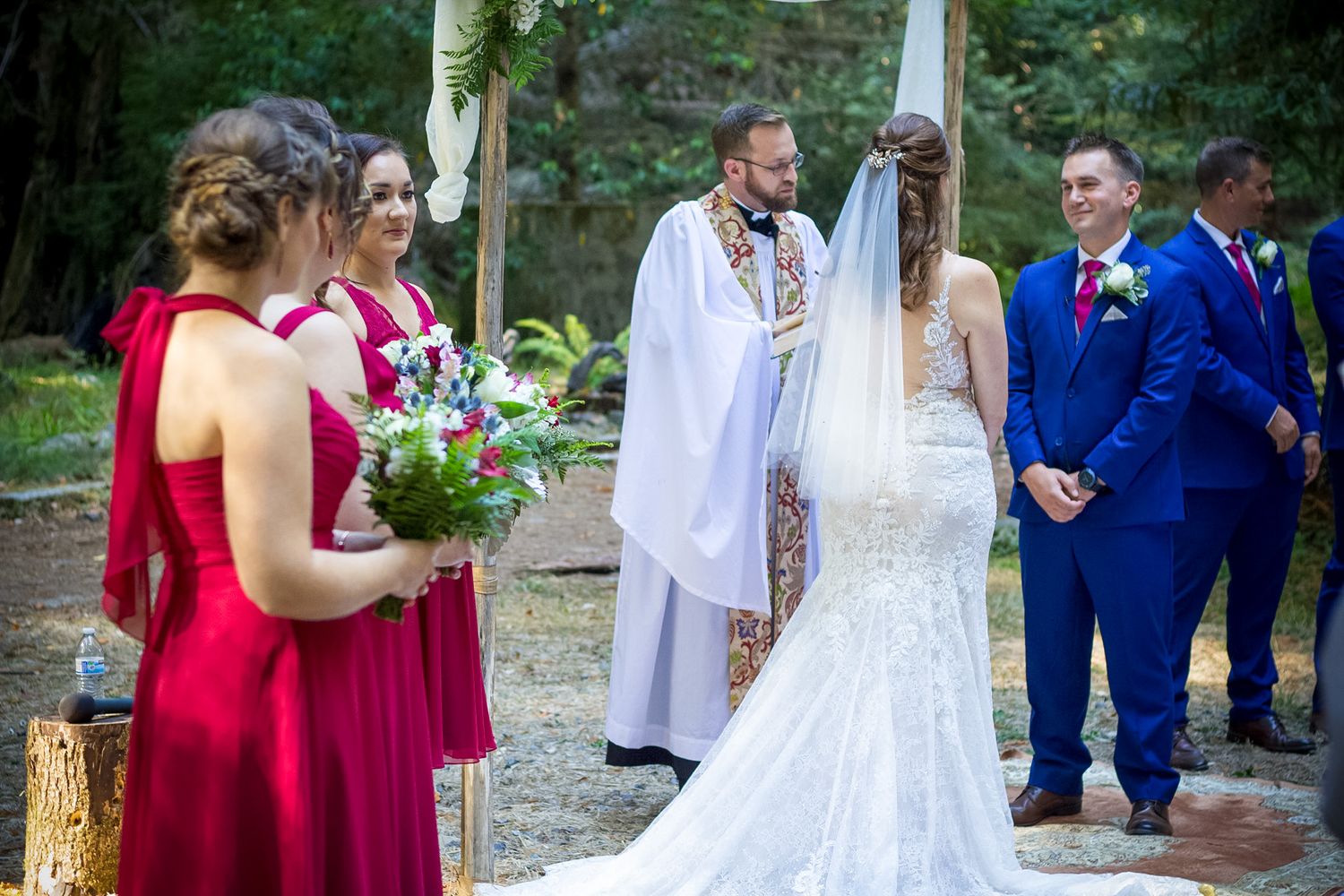A wedding ceremony in progress with attendants in red and blue attire gathered in a forest setting.