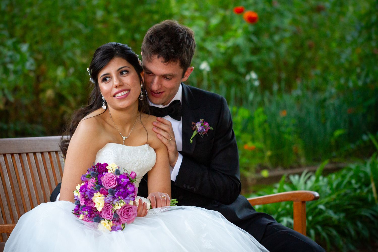 Romantic newlyweds share intimate moments in a lush garden setting with purple bouquet details.