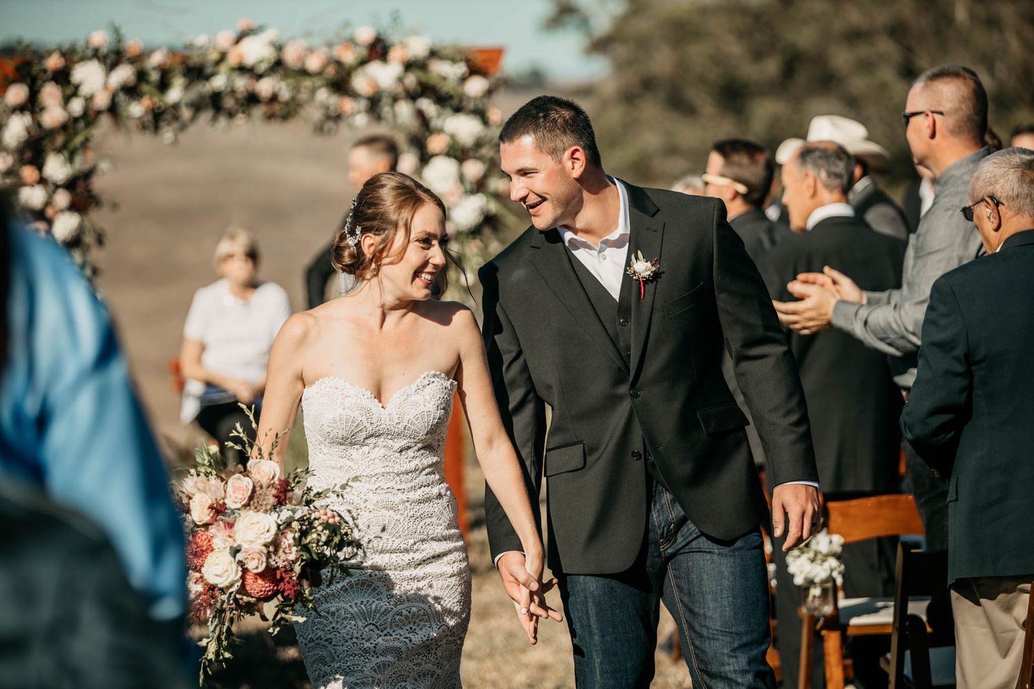 Newlyweds walking down aisle with floral arch and guests in background.