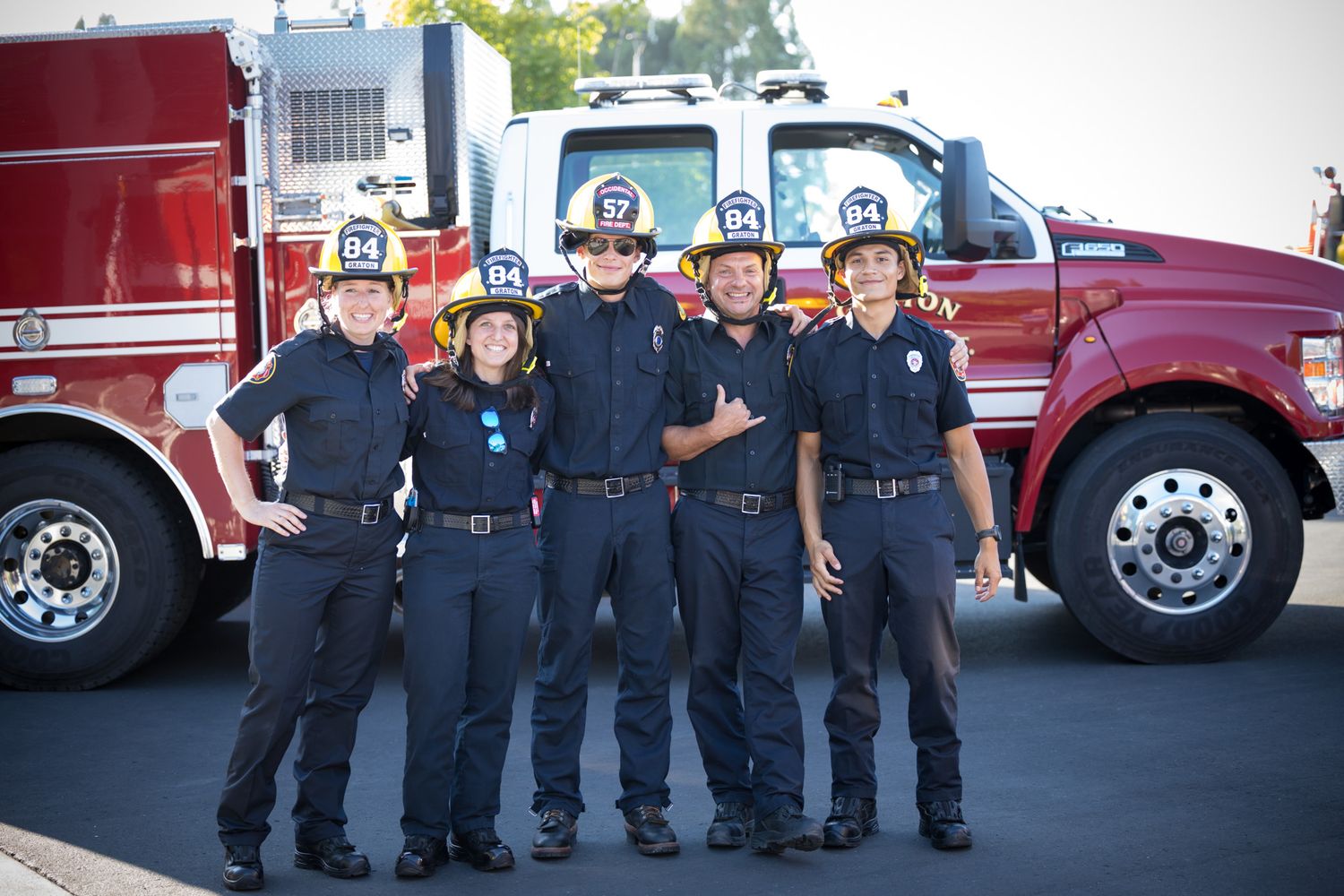 Group of firefighters lined up in front of red fire trucks in uniform.
