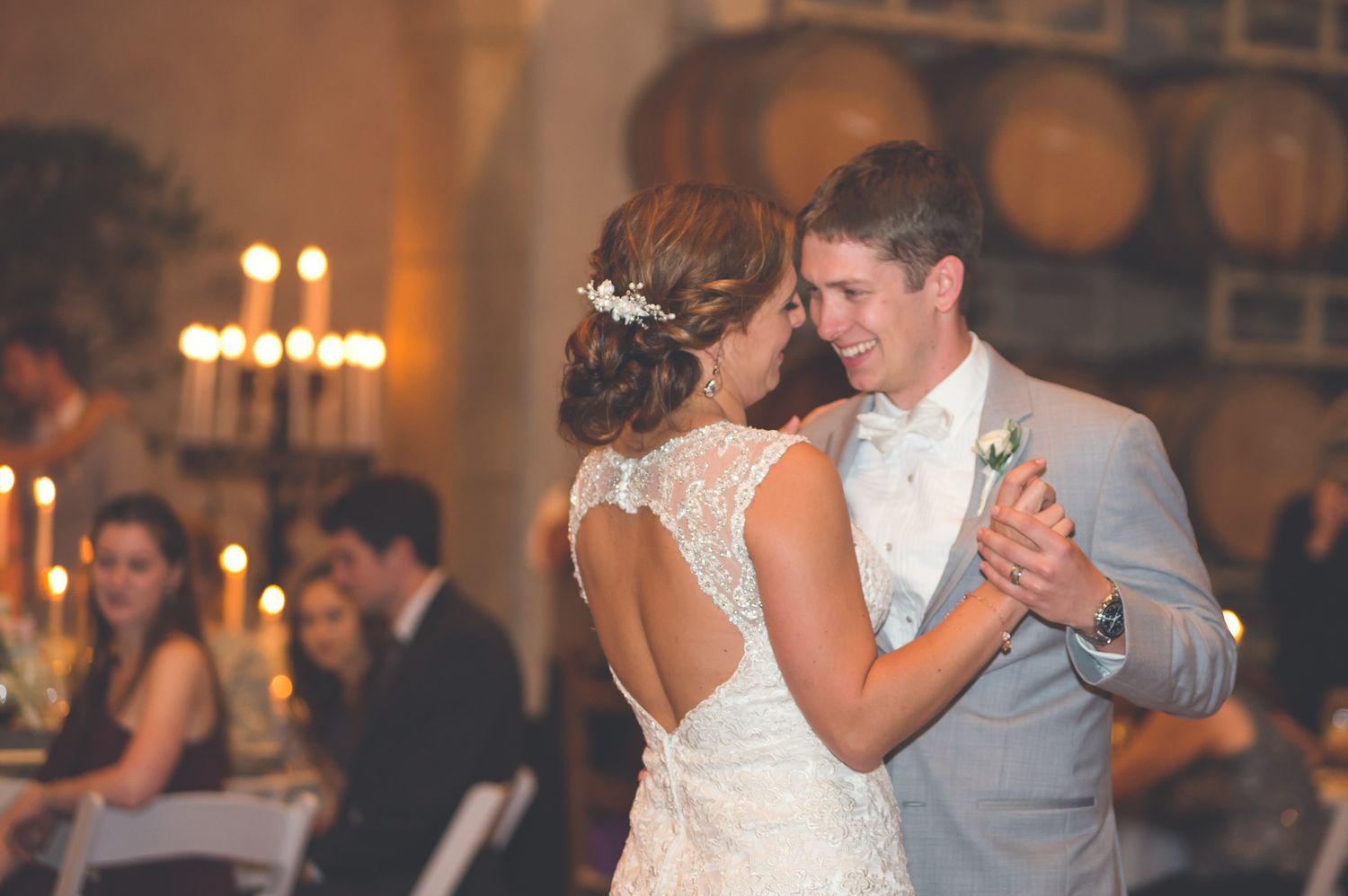 A couple shares their first dance at a romantic candlelit winery reception with wine barrels in the background.