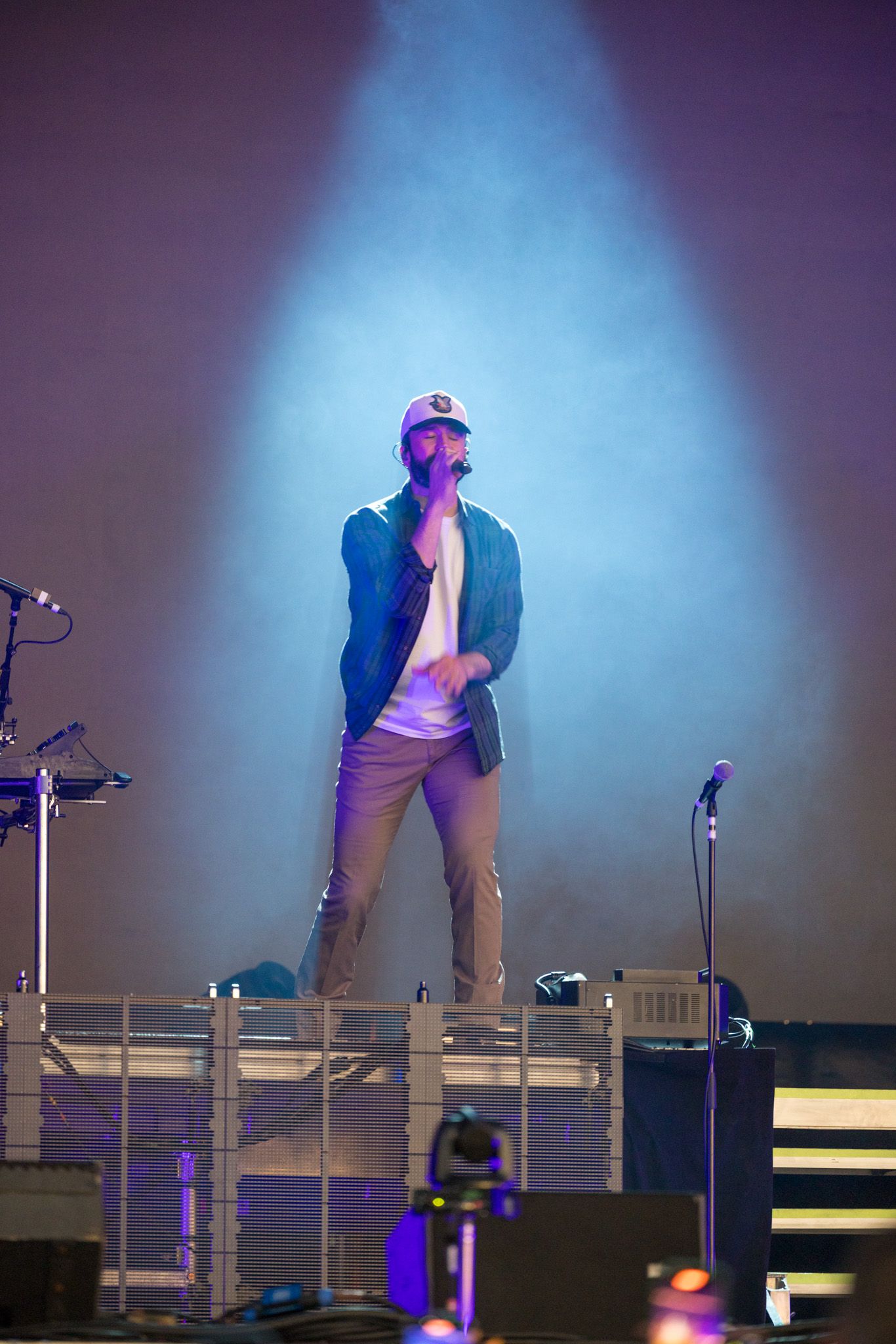 Performer in dark attire moves energetically on dimly lit stage with dramatic blue lighting and mic stands visible.