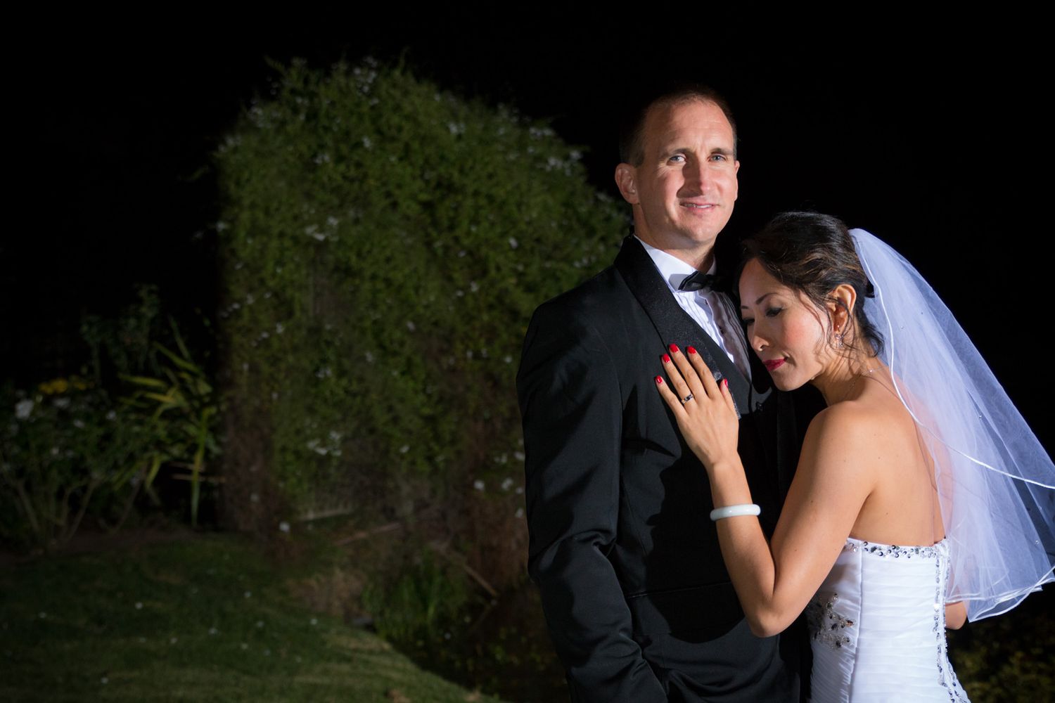 A couple in wedding attire poses together outdoors at night with trees in the background.