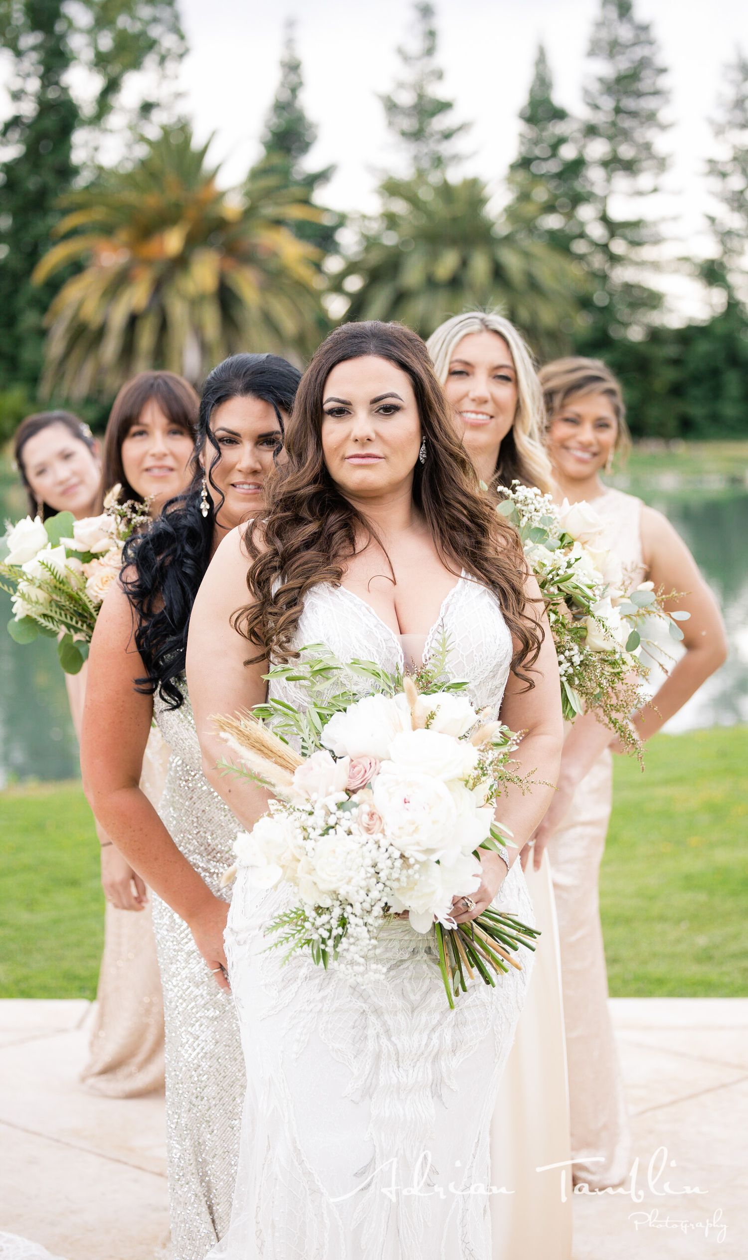 A bridal party in neutral dresses poses with bouquets by a lakeside garden.