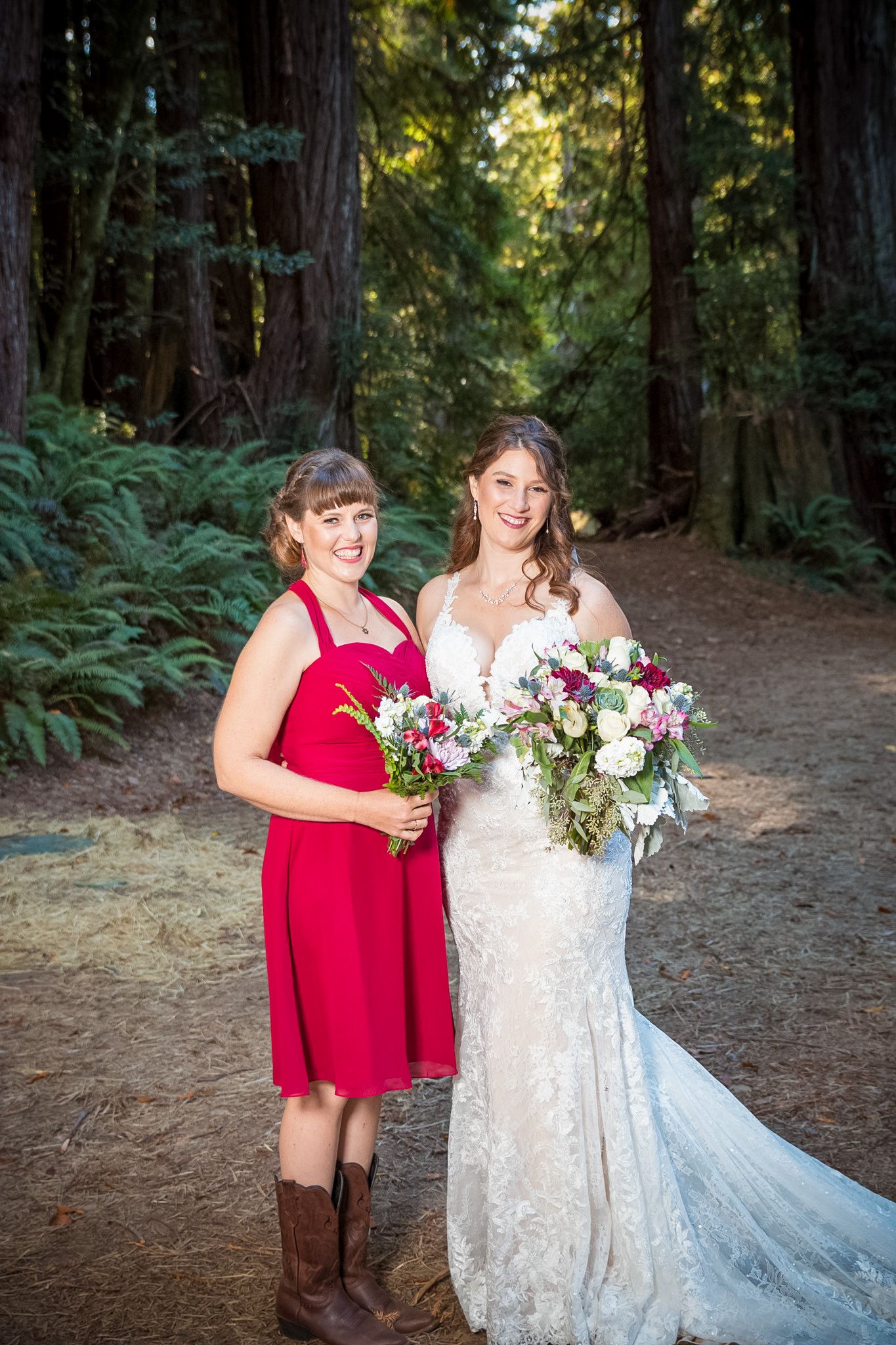 Bride in lace gown poses with bridesmaid wearing red dress and cowboy boots in forest setting.