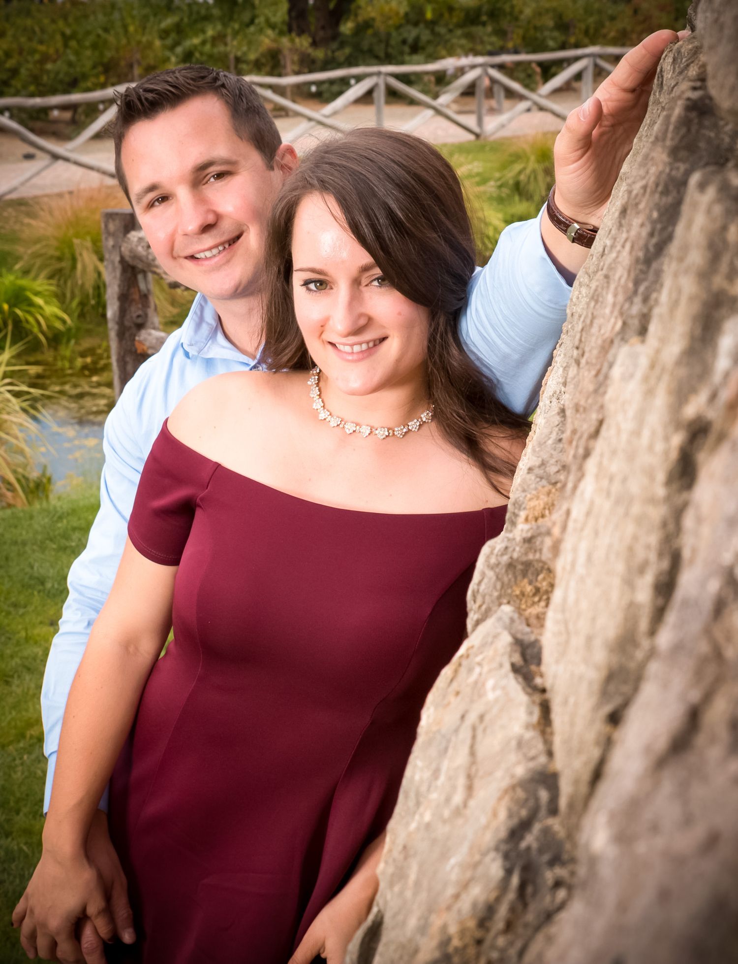 Couple smiles while posing next to a tree trunk in a garden setting.