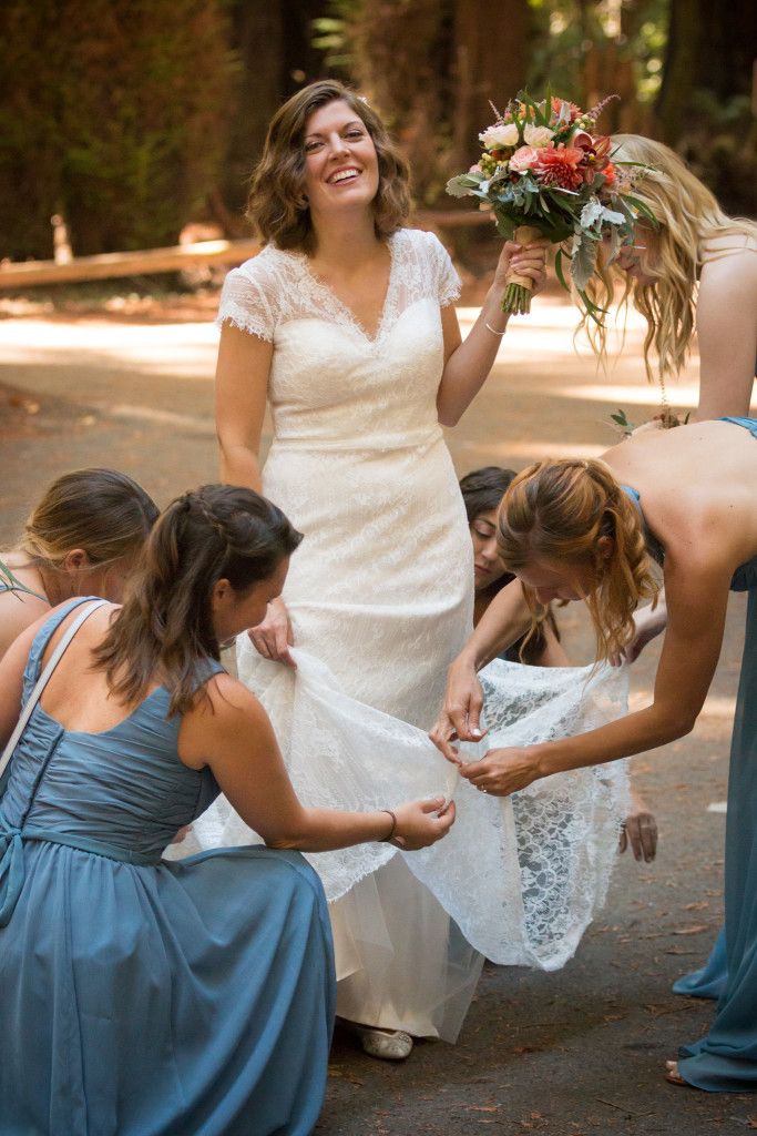 Bridesmaids help adjust the bride's lace dress hem during outdoor forest photoshoot.