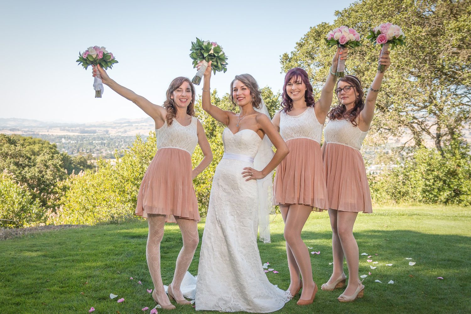 Bridesmaids in blush pink dresses stand with bride in white gown outdoors on a sunny hilltop lawn.