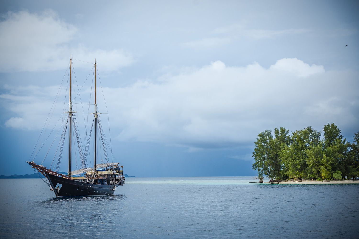 A traditional sailing vessel anchored in calm waters near a small forested island under cloudy skies.